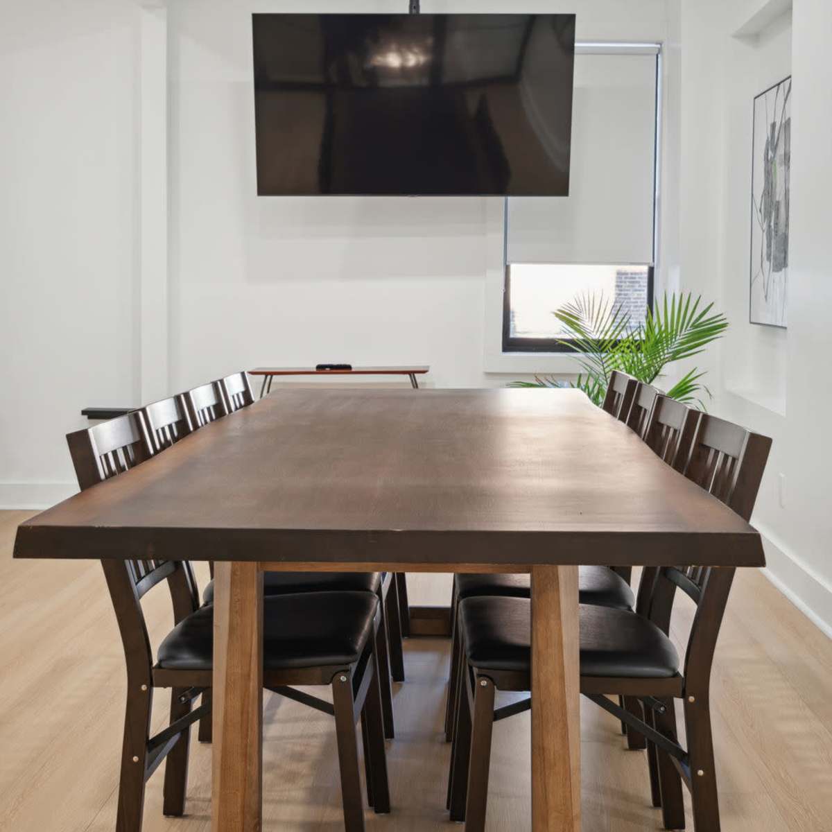 A large wooden dining table surrounded by eight black chairs is positioned in a bright room, with a wall-mounted TV above it and a plant visible in the corner.