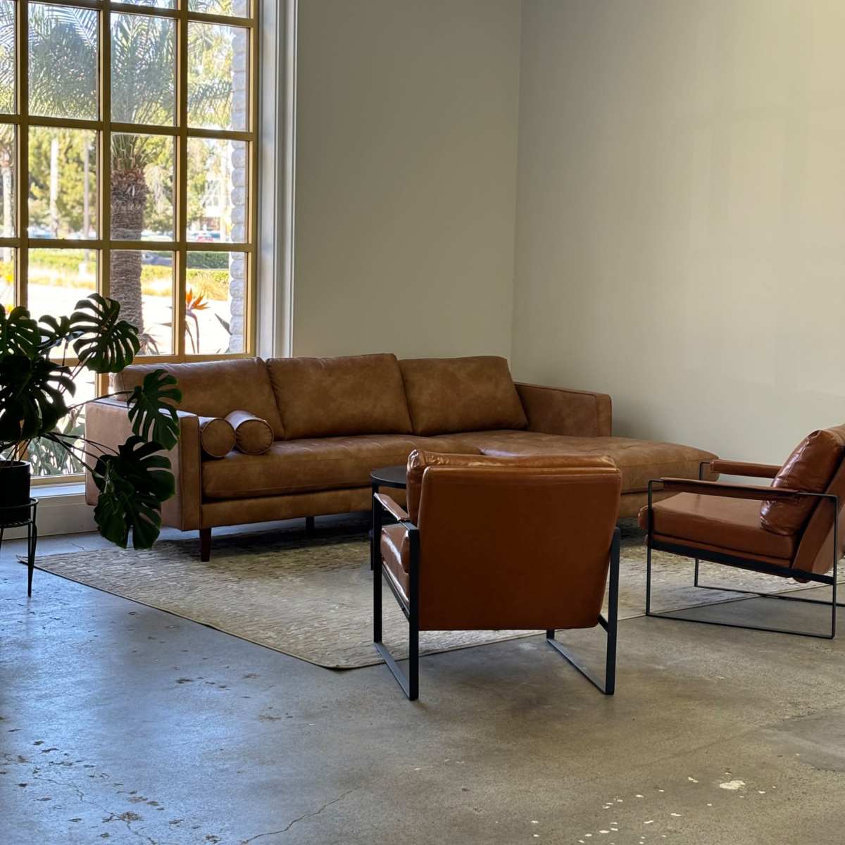A modern interior space featuring a brown leather sectional sofa, two brown leather chairs, a decorative plant, and a large window with palm trees visible outside.