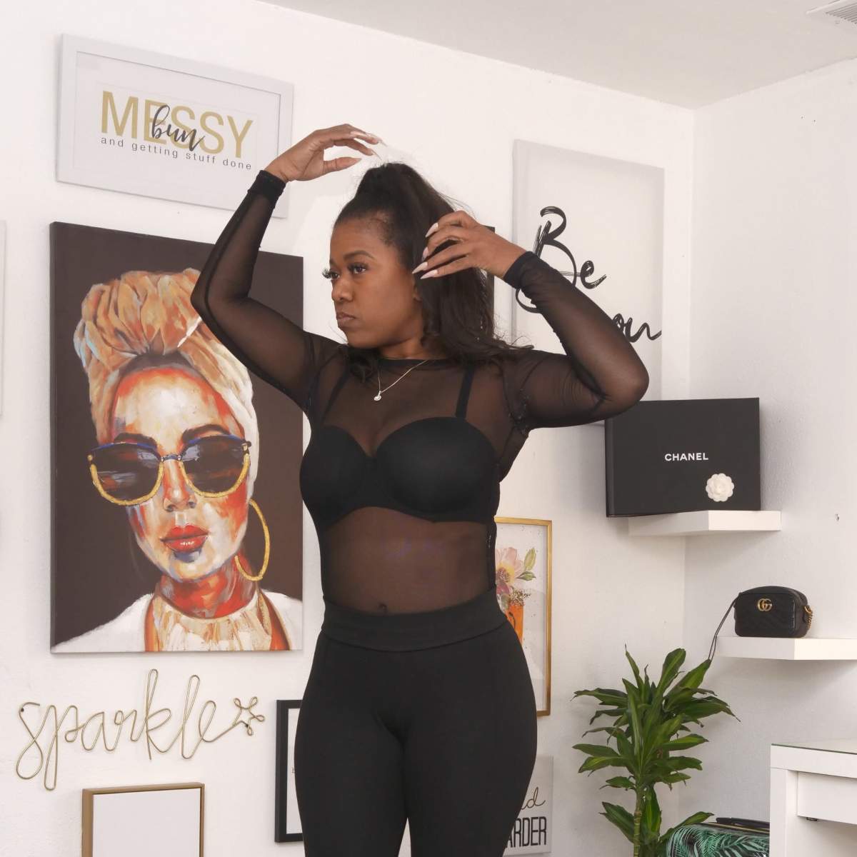 A woman in a sheer black top and leggings stands in a room decorated with artwork and motivational phrases.