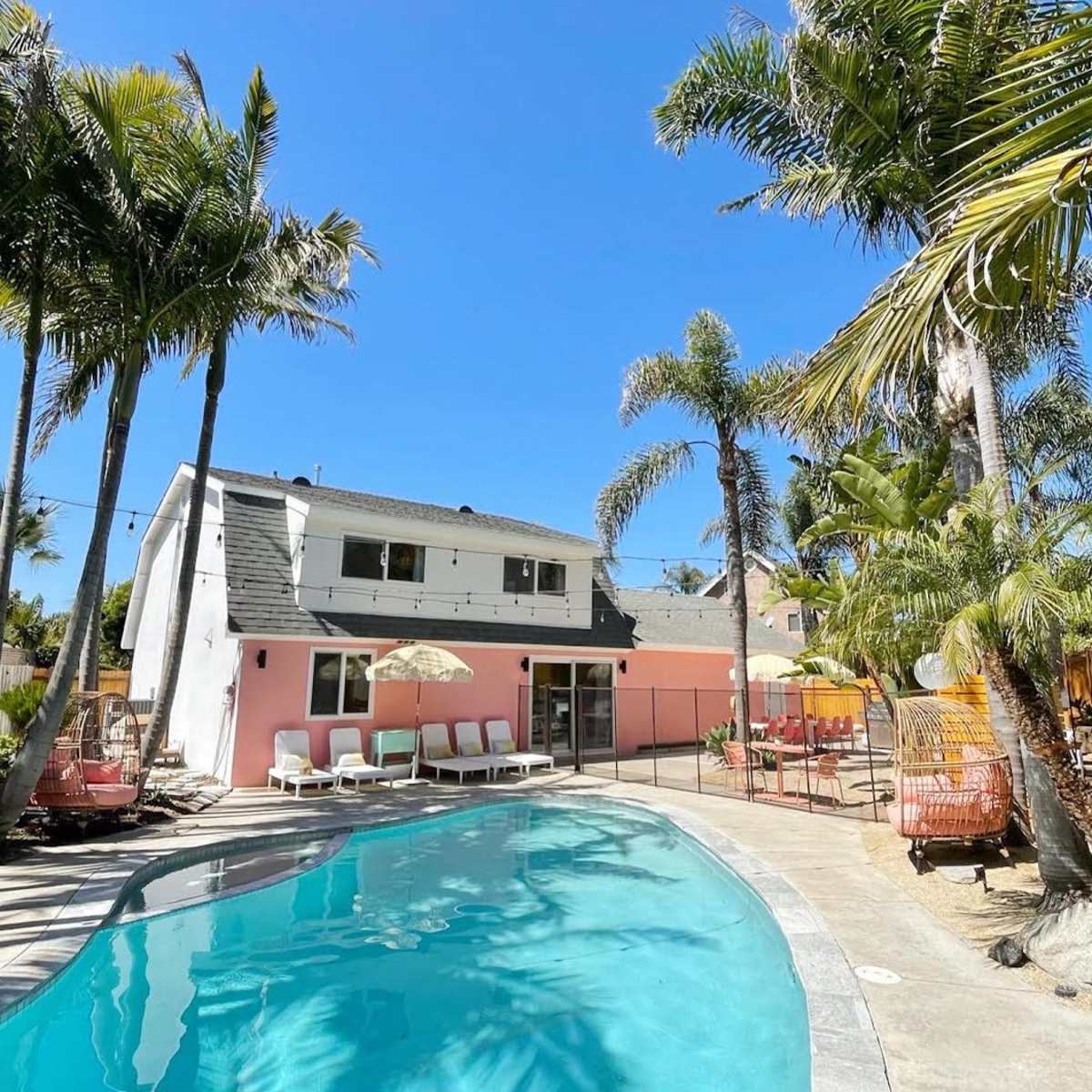 A swimming pool surrounded by palm trees and a pink house, with lounge chairs and an umbrella nearby.