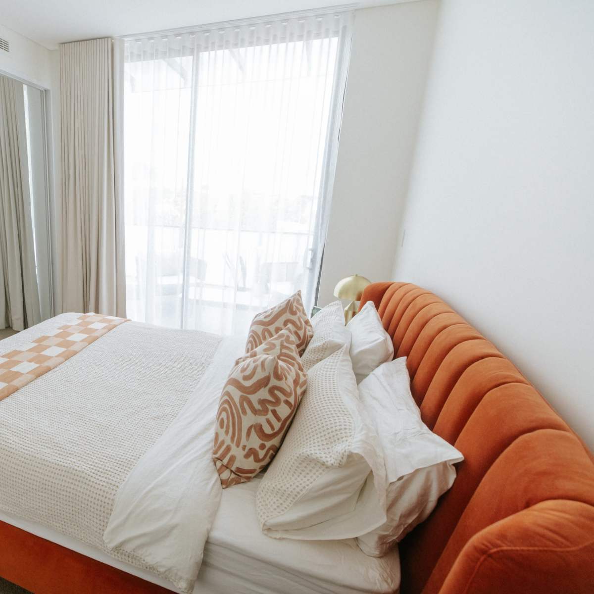 A well-lit bedroom featuring an orange upholstered bed with white bedding, decorative pillows, and a mirror reflecting part of the room.