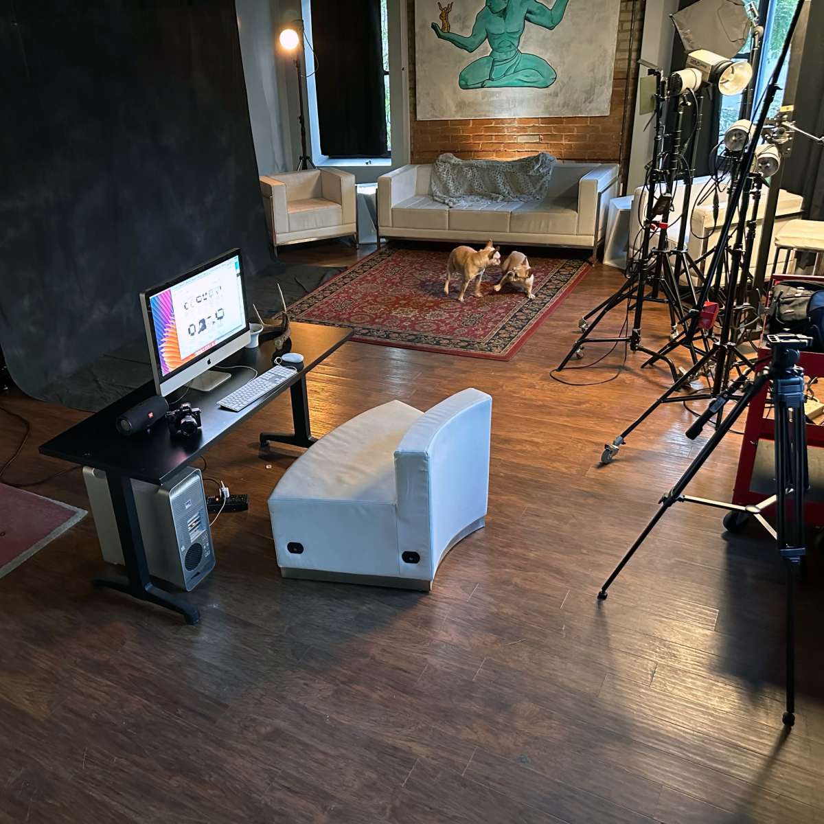 A studio space with a desk featuring a computer, a chair, and a backdrop alongside clothing racks, lighting equipment, and a couch.