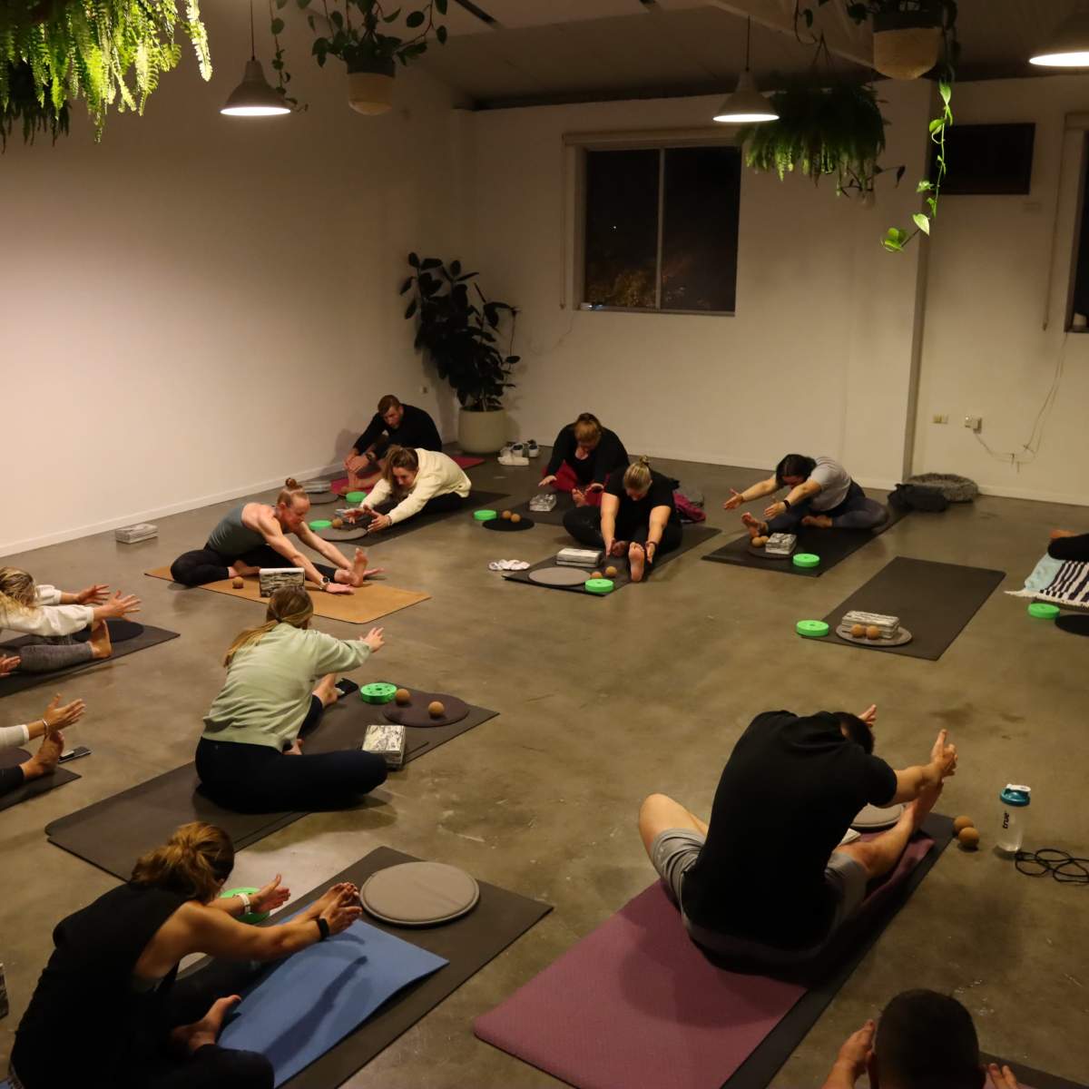 A group of people practice yoga in a spacious, dimly lit studio with mats and props scattered around.