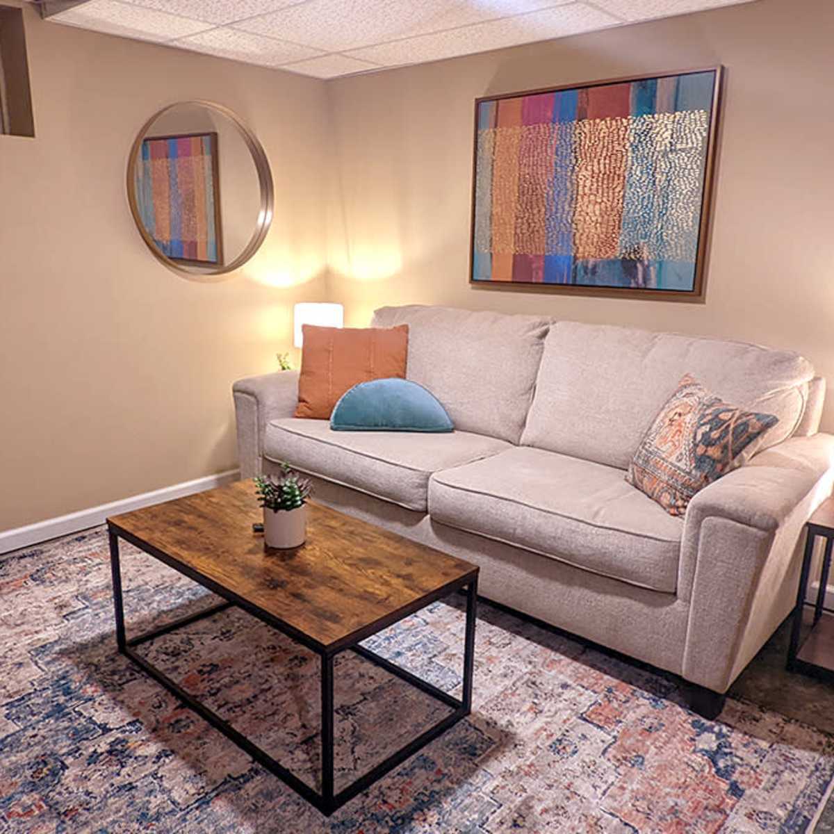 A small, neatly arranged living room with a light gray sofa, a wooden coffee table, a decorative mirror, and a colorful area rug.