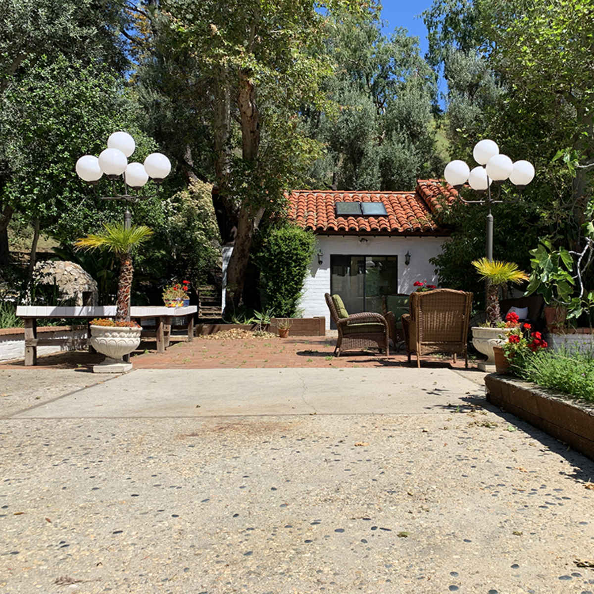 A spacious outdoor area featuring a mix of seating arrangements, decorative plants, and a house with a red tile roof, surrounded by trees.