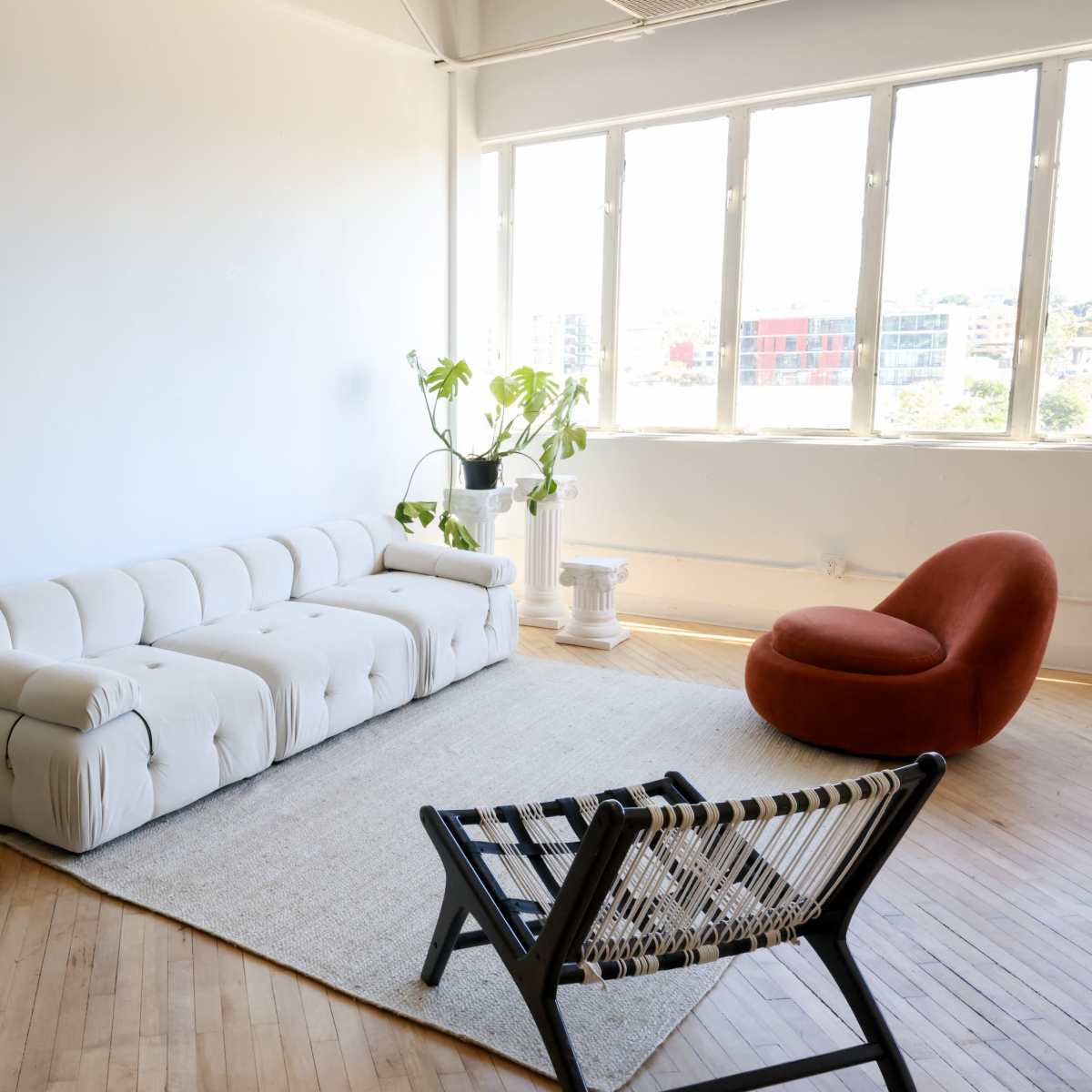 A minimalist living space featuring a beige couch, a red chair, a large mirror, and a plant, all arranged in front of a row of windows.