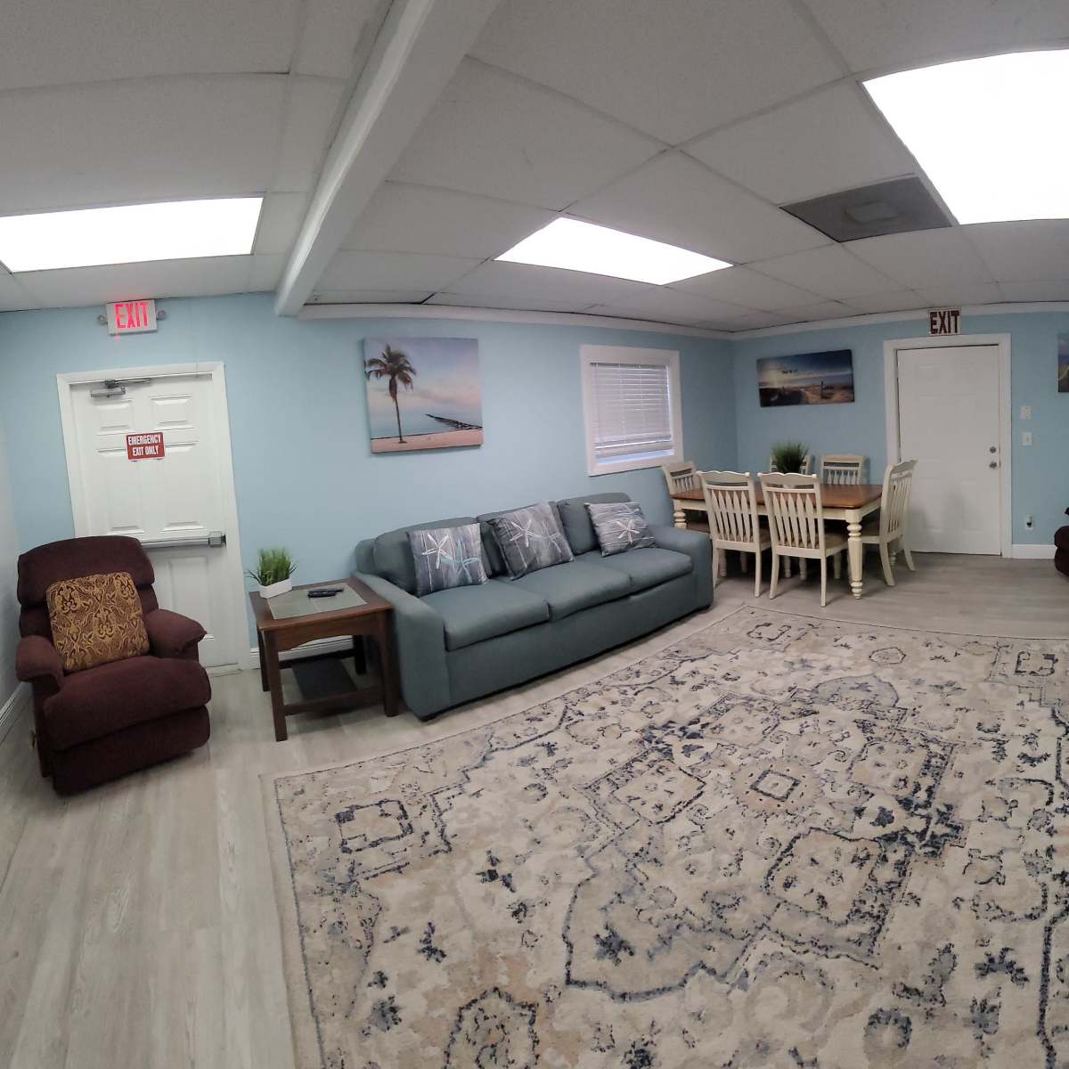 The room features a blue wall color, a dark sofa, an area rug with a pattern, a dining table with chairs, and a large television mounted on the wall.
