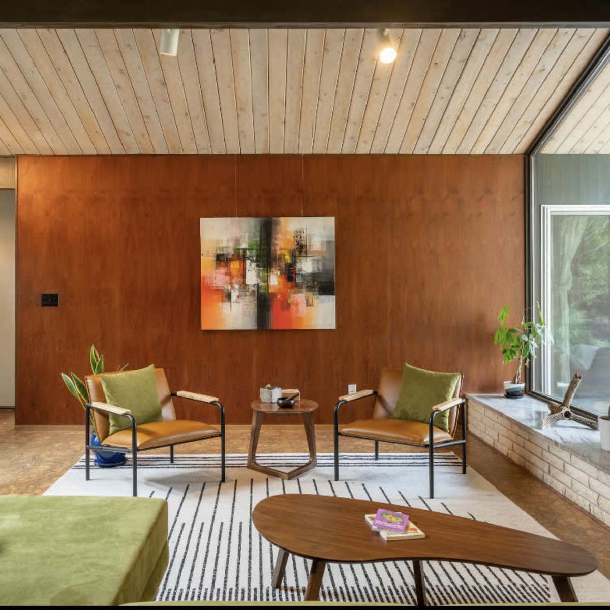 A modern living room featuring two green chairs and a sofa, a wooden coffee table, and a large artwork on a brown wall.
