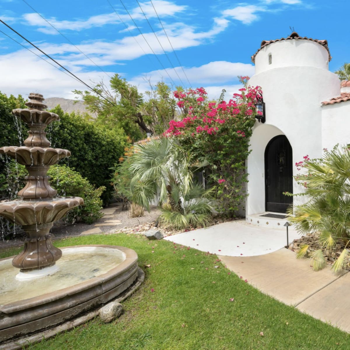 A white stucco house with a decorative entrance features a prominent fountain and colorful landscaping.