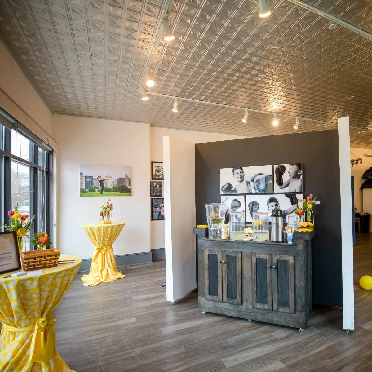 The interior of a brightly lit event space features a serving station against a black wall, surrounded by decorated tables with yellow tablecloths.