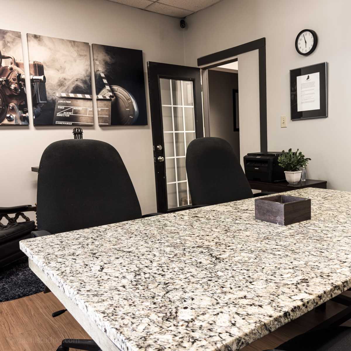 A conference room features a granite table surrounded by black chairs, with framed artwork and a clock on the walls.