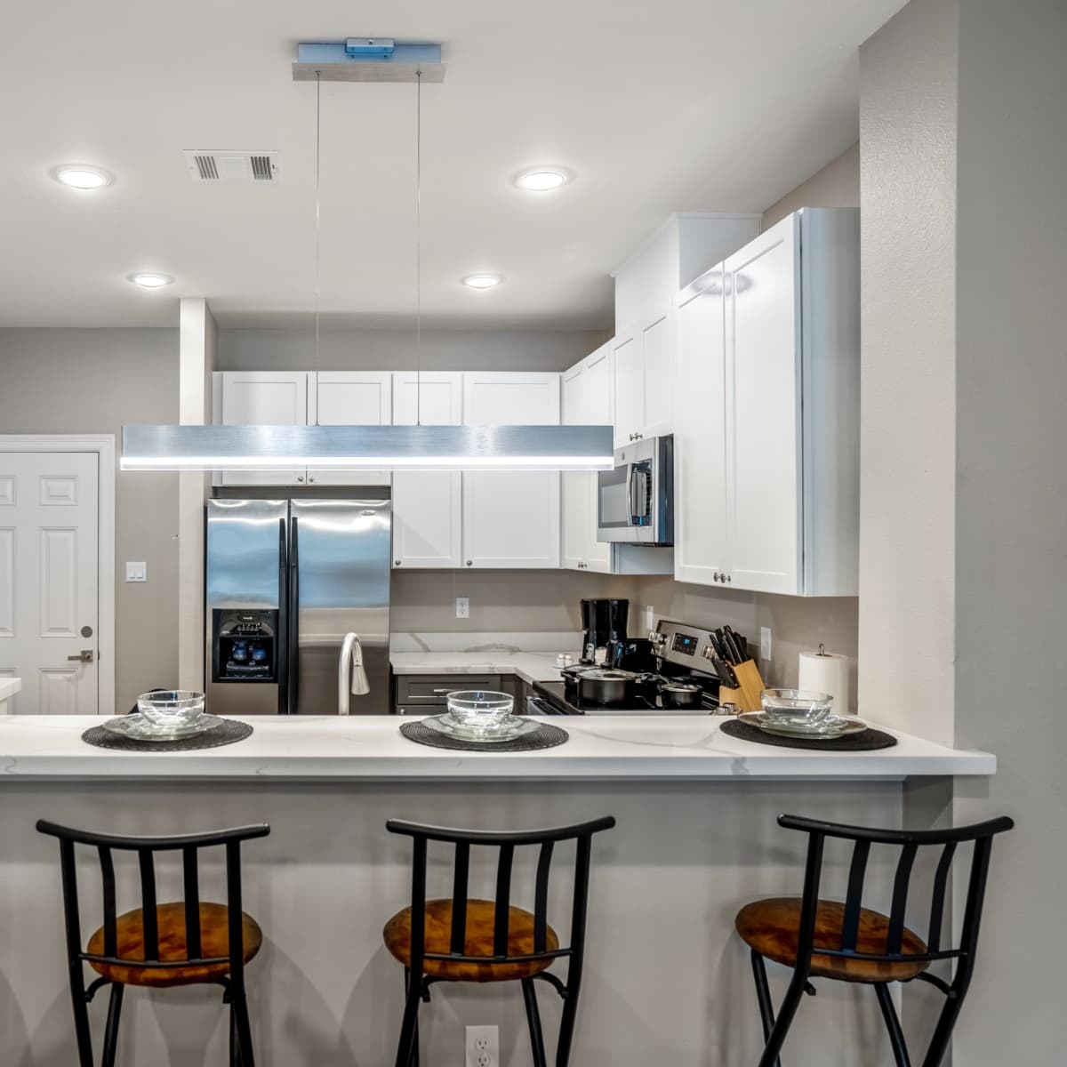 A modern kitchen features white cabinetry, stainless steel appliances, and a bar with four black chairs.