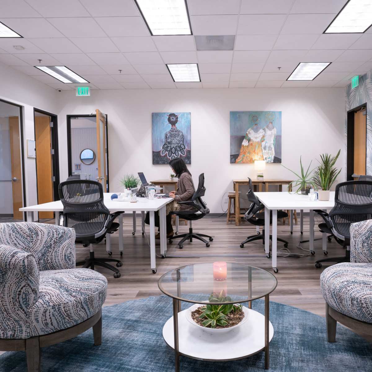 A modern office space features two seating areas with patterned chairs, a round table, and individual workstations equipped with computers against a backdrop of artwork and decorative plants.