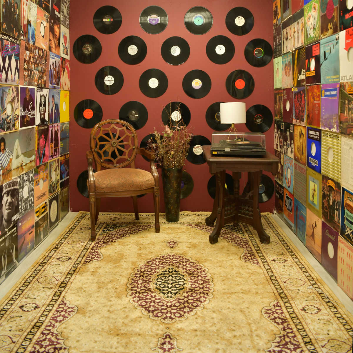 A vintage room features a chair and a record player against a backdrop of colorful vinyl records mounted on the walls.
