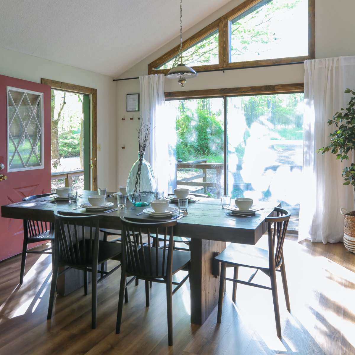 A dining area with a wooden table set for a meal, surrounded by black chairs, and large windows allowing natural light to fill the space.