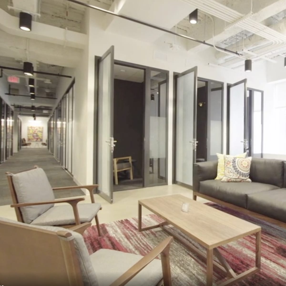 A modern office space with a small seating area featuring a sofa, armchairs, and a coffee table, complemented by a hallway of glass-walled meeting rooms.