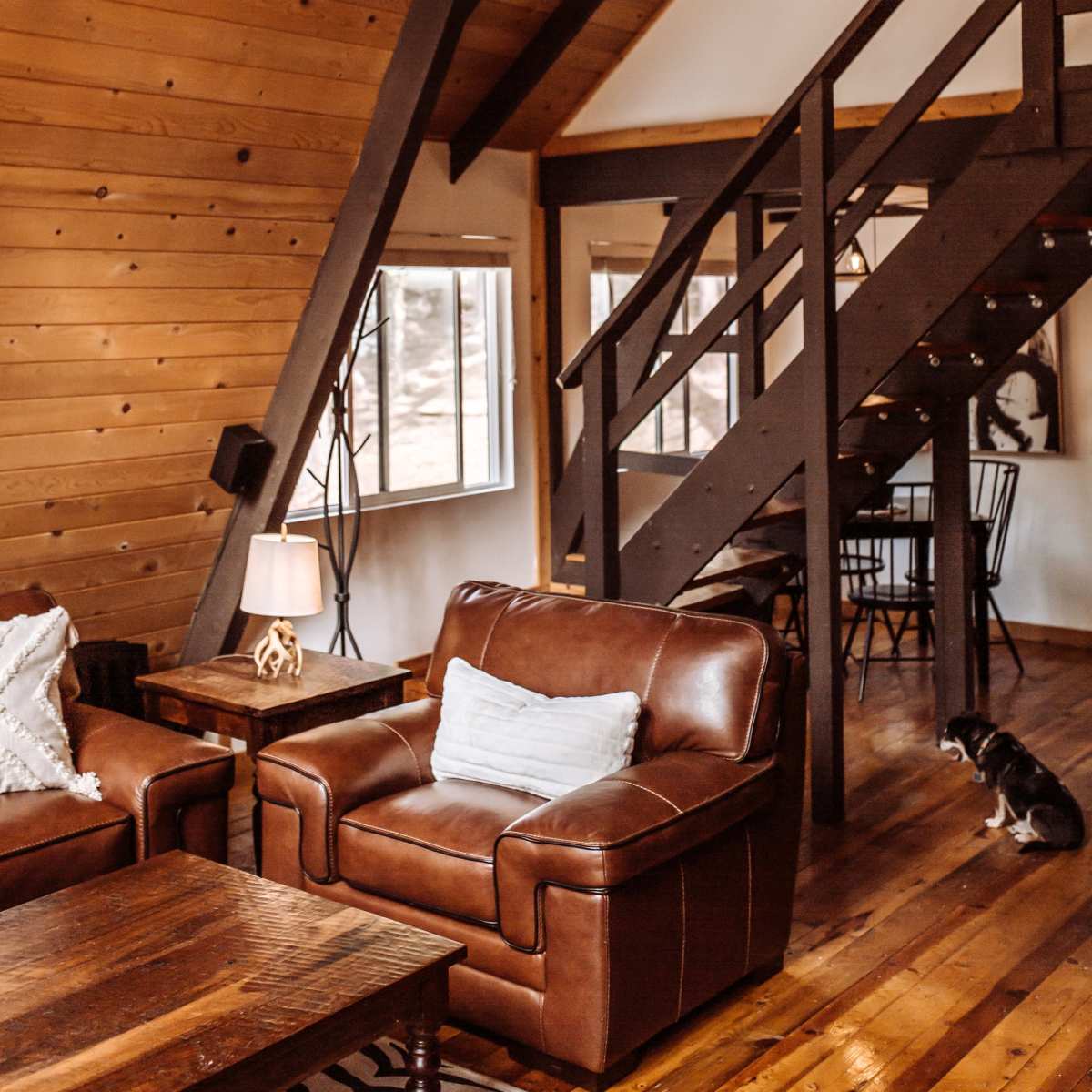 A rustic living room with a leather couch, wooden flooring, and a large chandelier made of antlers, alongside a staircase leading to the upper level.