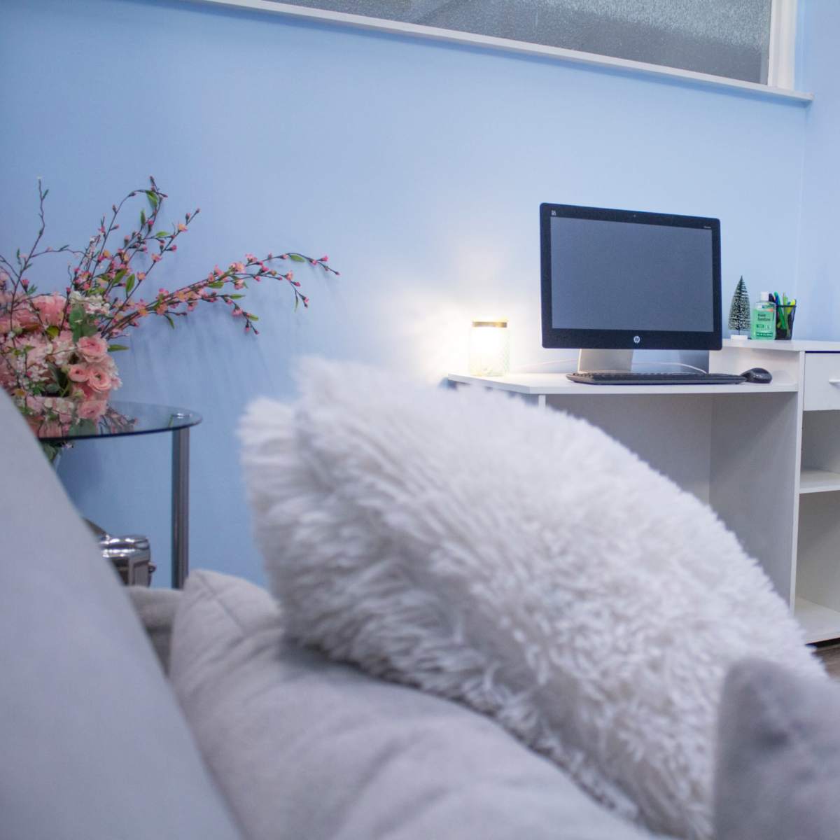 A light blue room with a computer on a white desk, a decorative plant, and a floral arrangement next to a gray sofa.