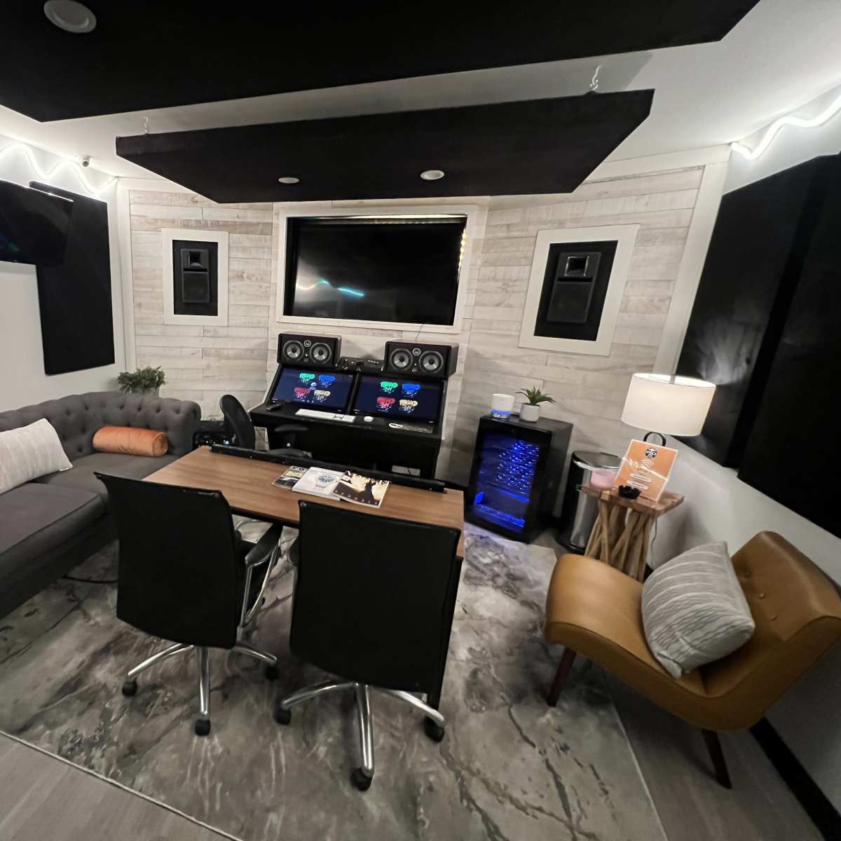 A modern recording studio with a gray sofa, a wooden table with two black chairs, and sound equipment mounted on the walls, all set against a light-colored wood panel background.