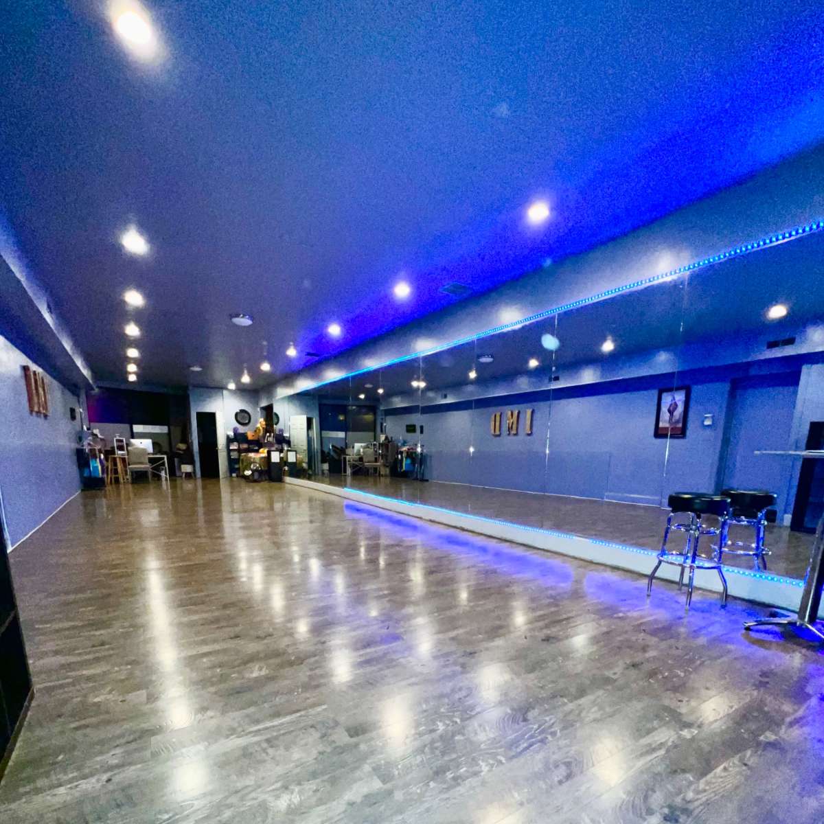 A spacious dance studio with wooden flooring, blue-lit walls, and mirrors reflecting the room's interior.