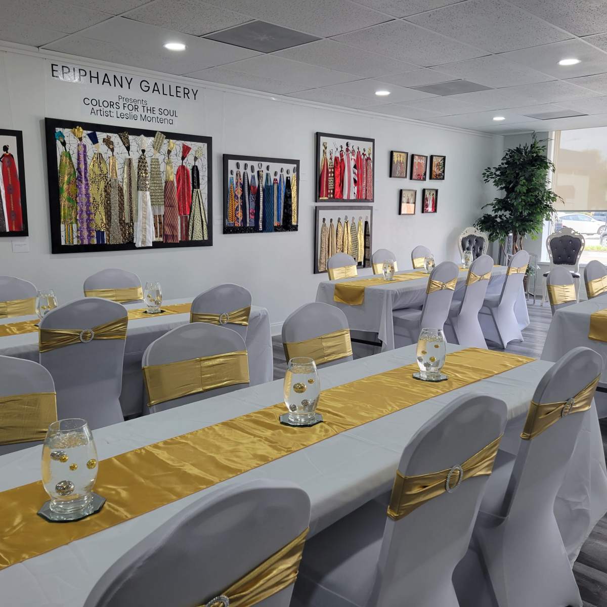 A neatly arranged banquet room with white tables adorned with gold table runners and decorated with glass vases in front of framed artworks on the walls.