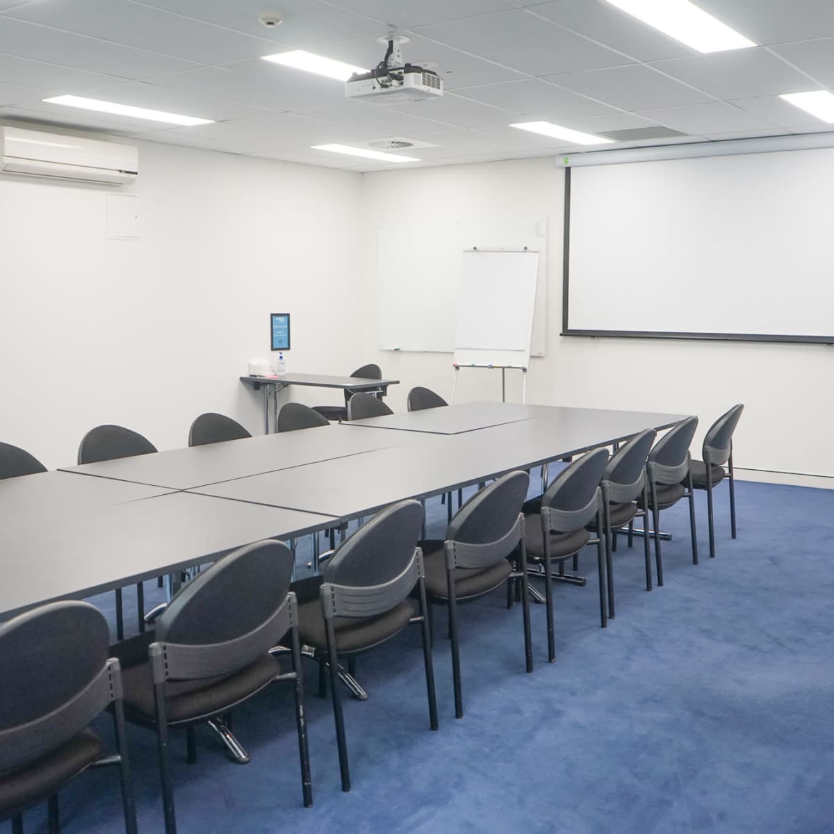 A conference room is set up with a long table and chairs, featuring a projector screen and whiteboards at the front.