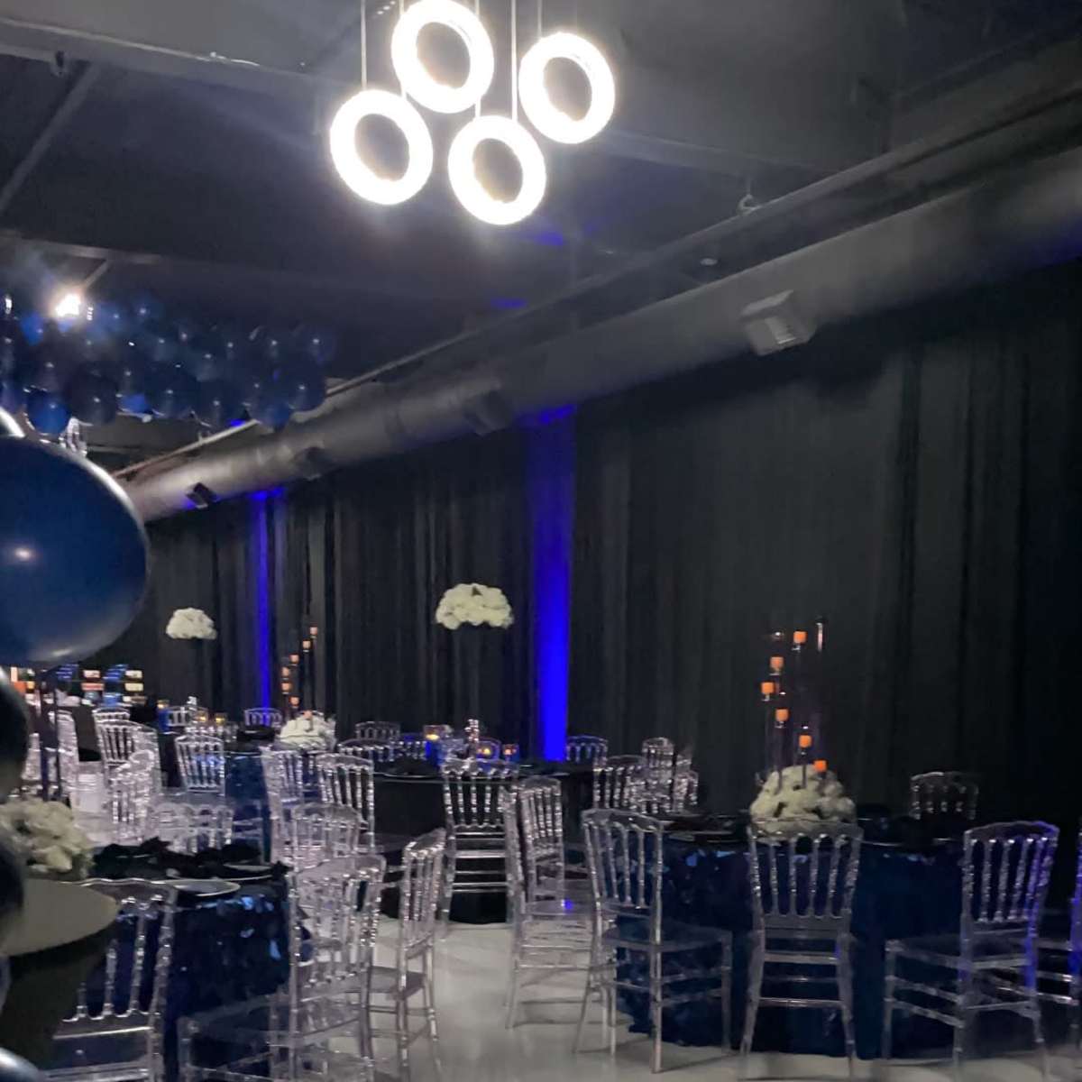 A decorated event space features elegant tables with dark tablecloths, clear chairs, and blue balloons overhead.