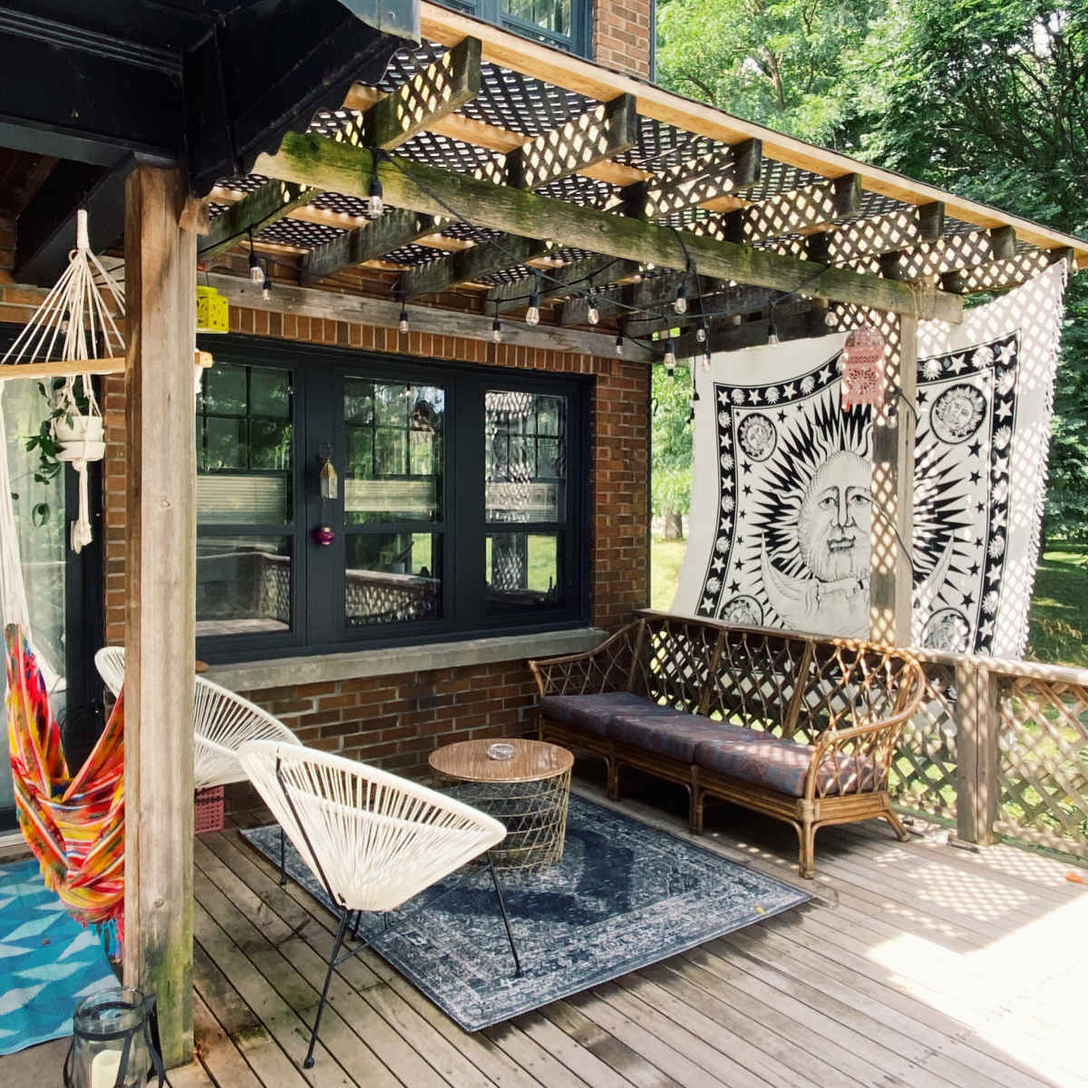 A wooden deck attached to a brick house, featuring a hammock, a rattan sofa, a round table, and a large tapestry hanging on the wall.