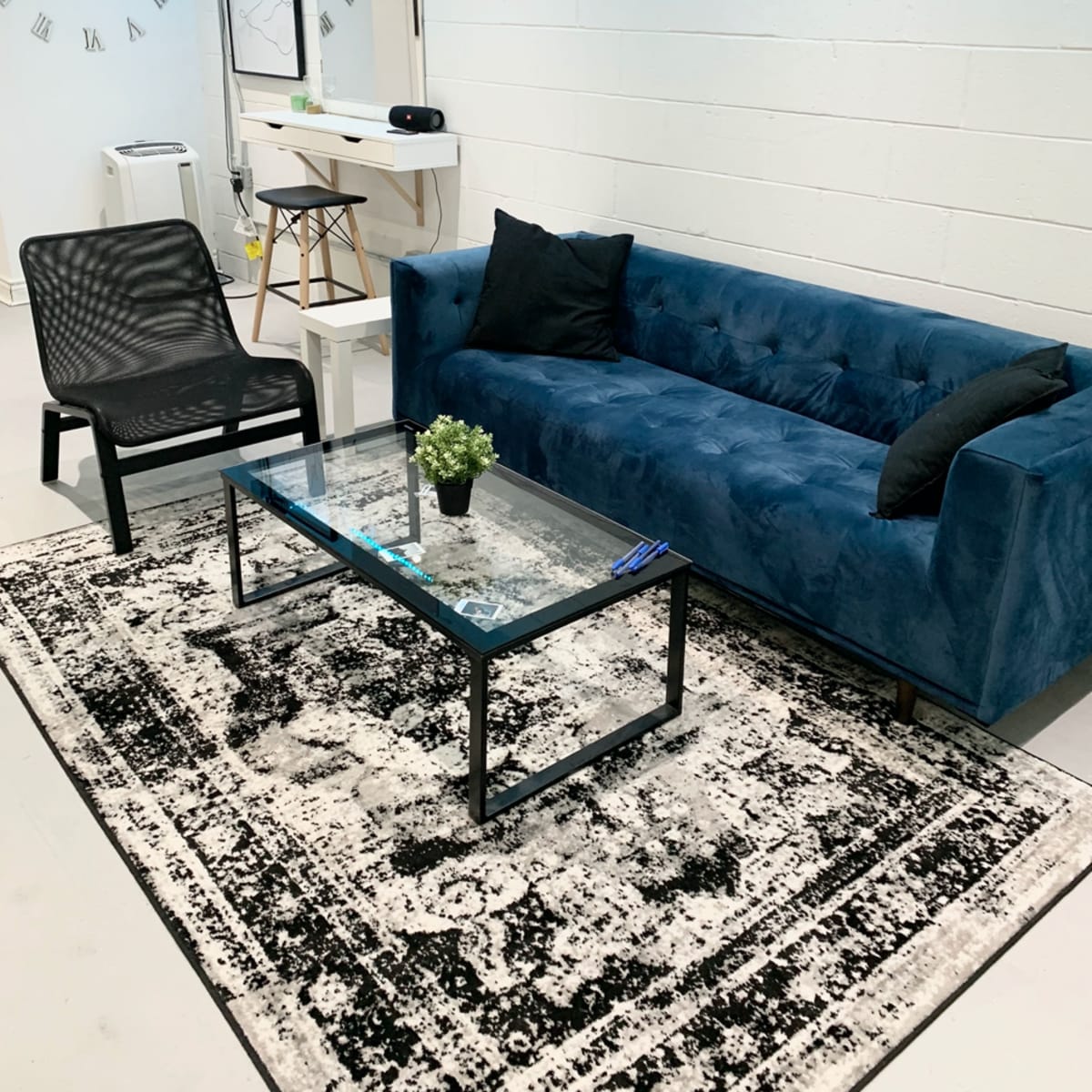 A modern living space featuring a blue velvet sofa, a glass coffee table with a small plant, and a black chair on a patterned rug.