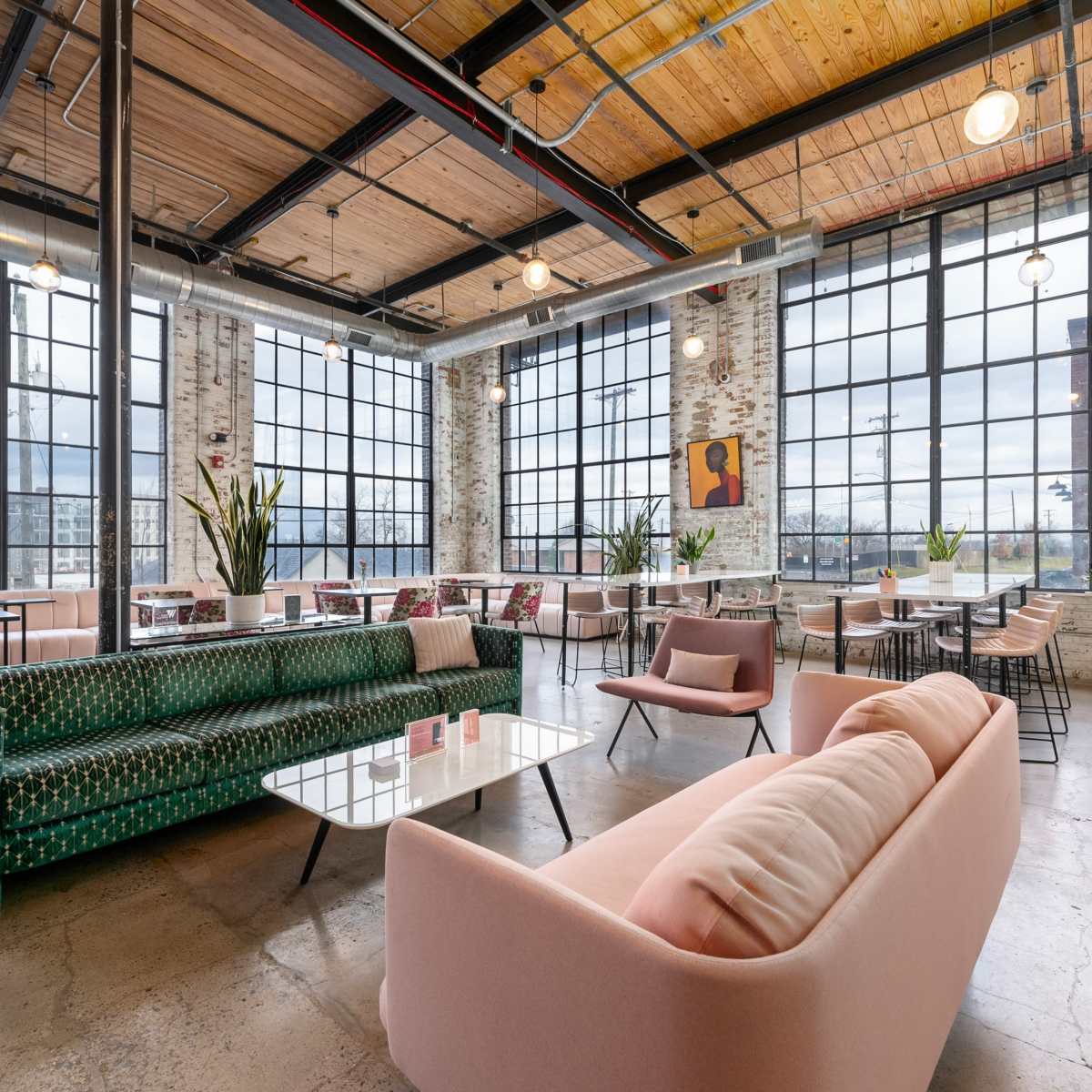 A modern café with large windows, exposed brick walls, and a mix of green and pink upholstered seating arranged around tables.