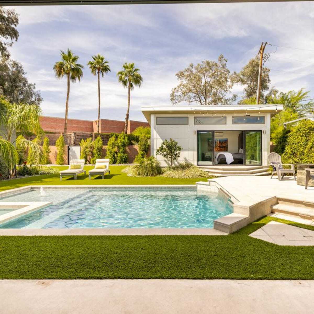 A modern backyard with a pool, lounge chairs, and palm trees, situated beside a contemporary house.