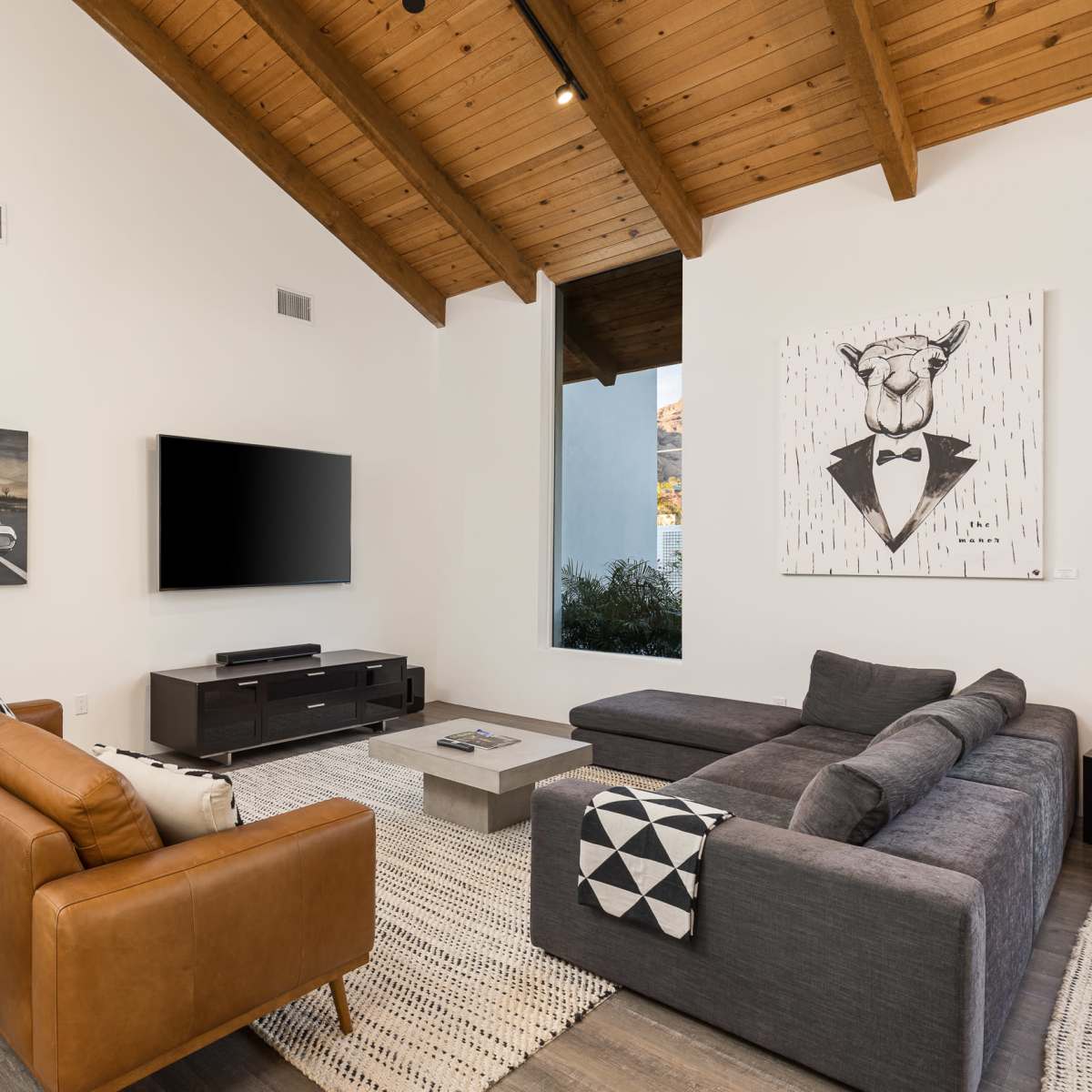 A modern living room features a large sectional sofa, a leather armchair, a flat-screen TV, and artwork on the walls, with vaulted wooden ceilings and natural light coming through a window.