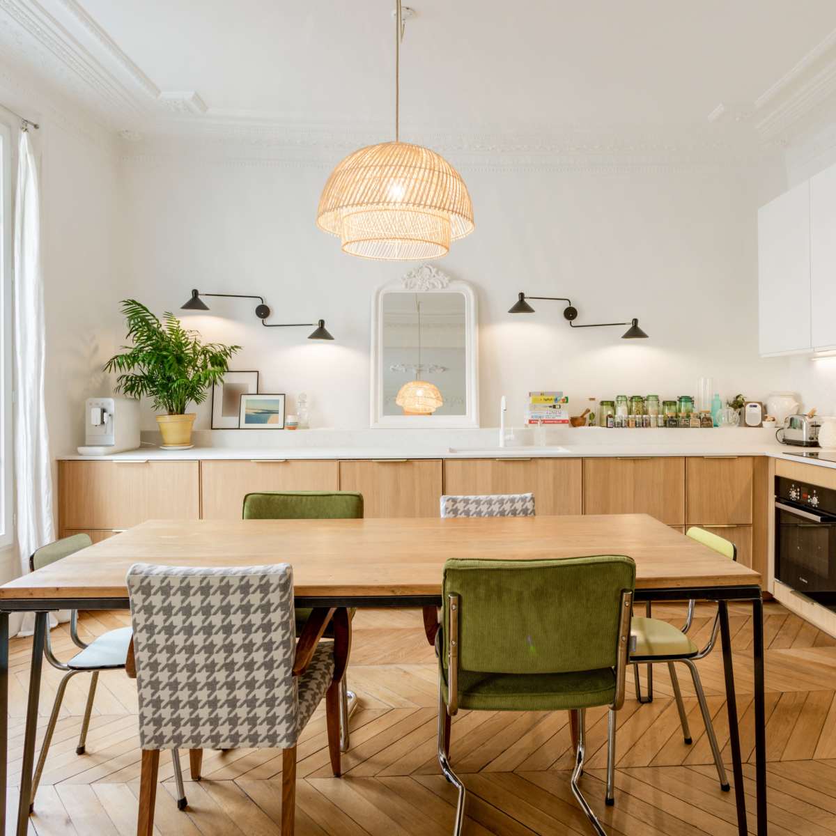 A modern kitchen features a wooden dining table surrounded by various chairs, with white cabinets and a large window allowing natural light to enter.