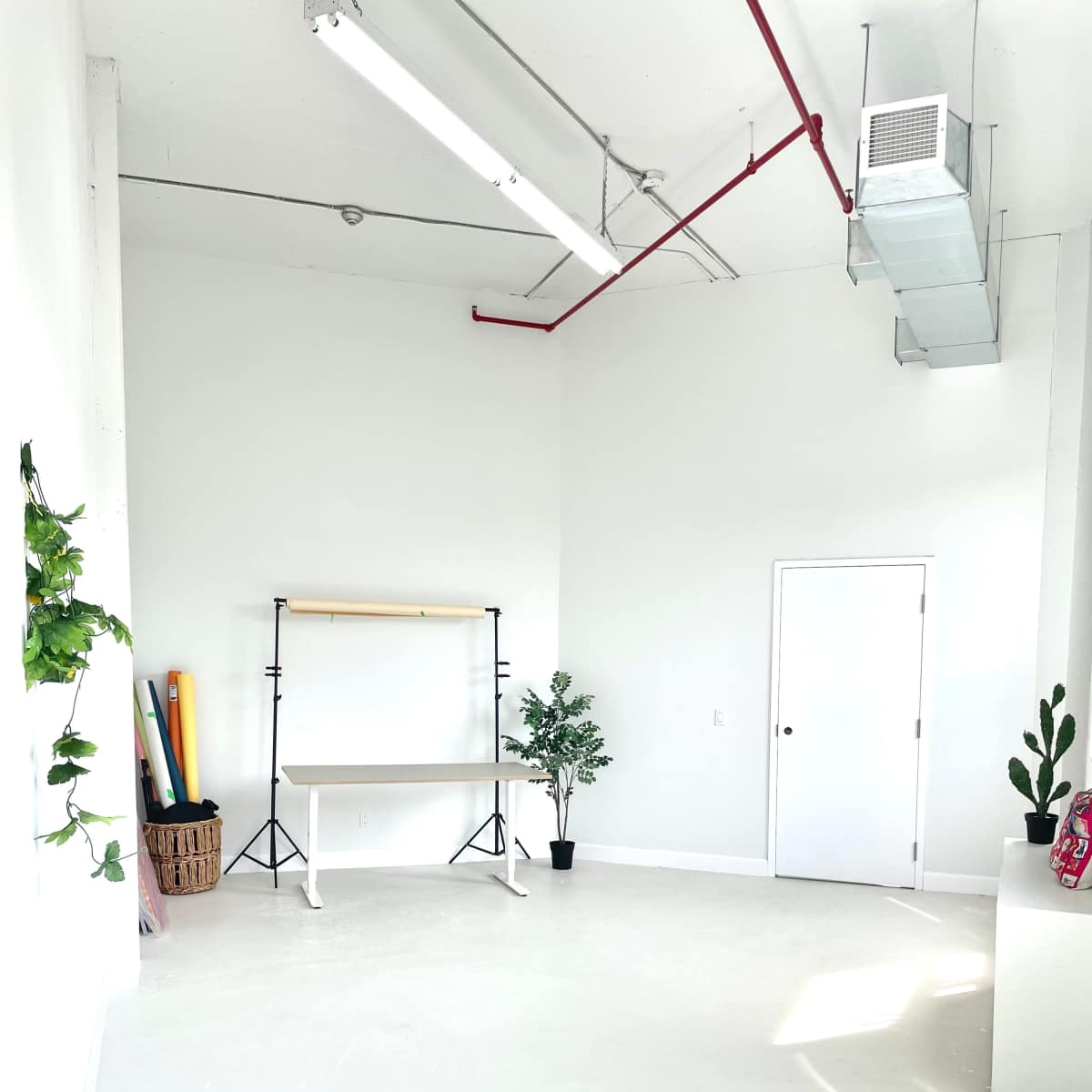 A spacious, bright studio with white walls, a table on a light-colored floor, and minimalist decor including plants and rolls of colored materials.