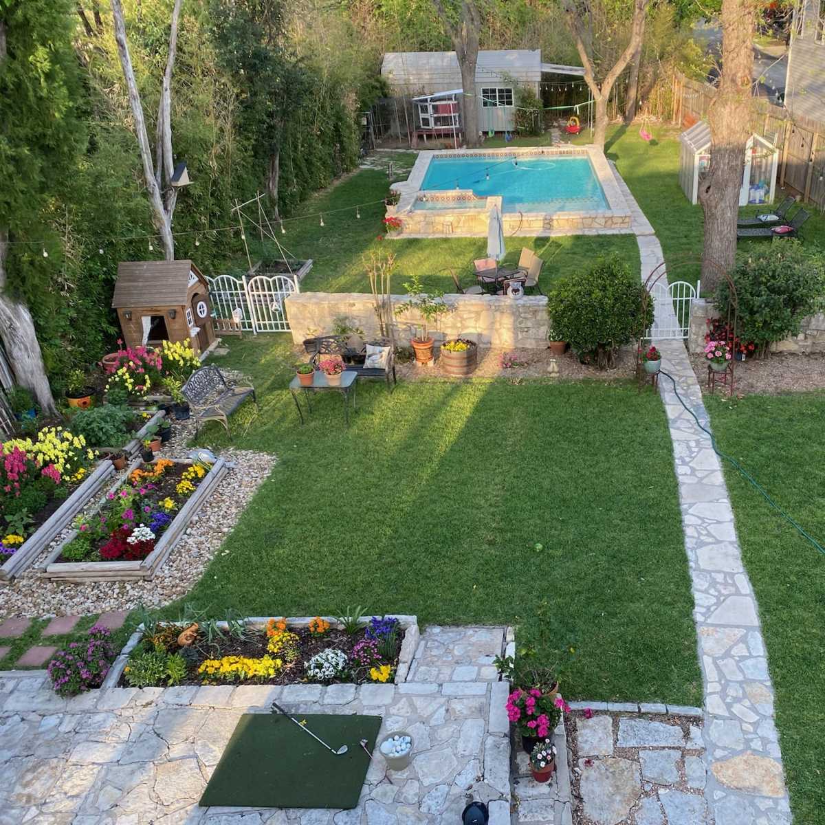 A landscaped backyard featuring a swimming pool, garden beds with colorful flowers, a stone path, and a children's play structure.