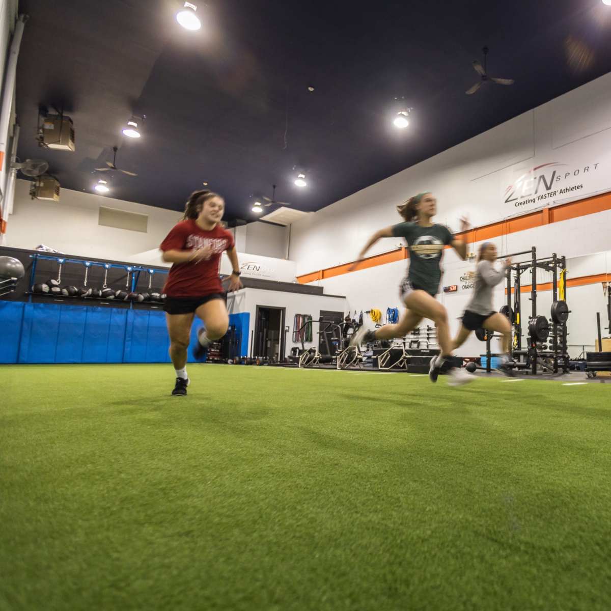 People are running on a turf surface inside a training facility equipped with weightlifting racks and exercise equipment.