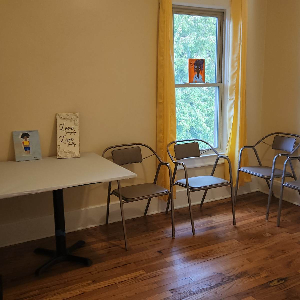 A small, empty room features a table and four metal chairs arranged around it, with yellow curtains framing a window.