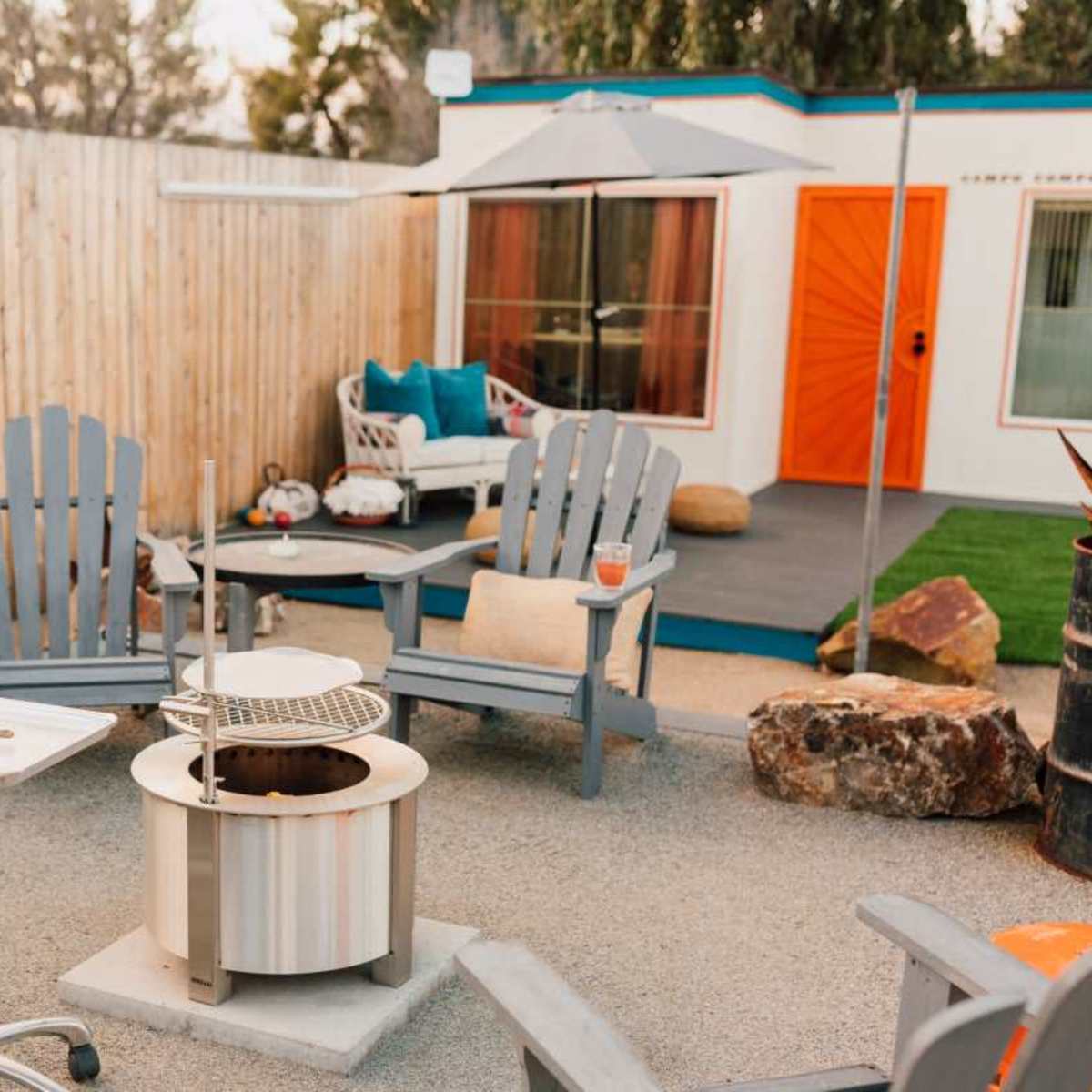 An outdoor seating area with several gray Adirondack chairs arranged around a circular fire pit, with a table and a small patio set in the background.