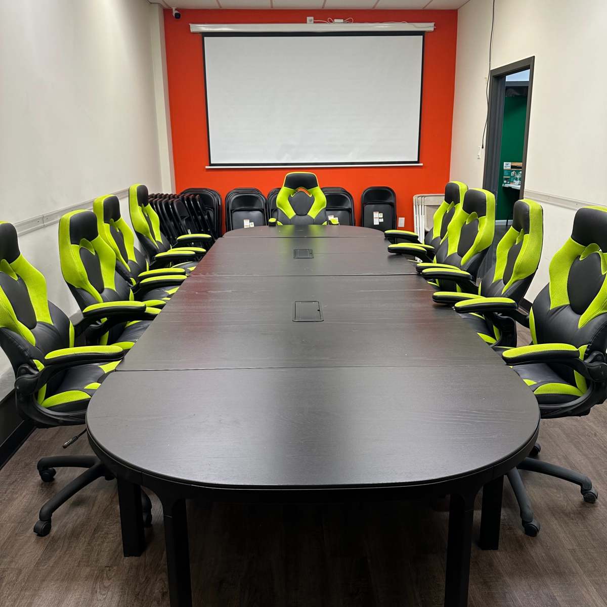 A rectangular conference table is surrounded by multiple black and green ergonomic chairs in a brightly lit meeting room with an empty projector screen on the wall.