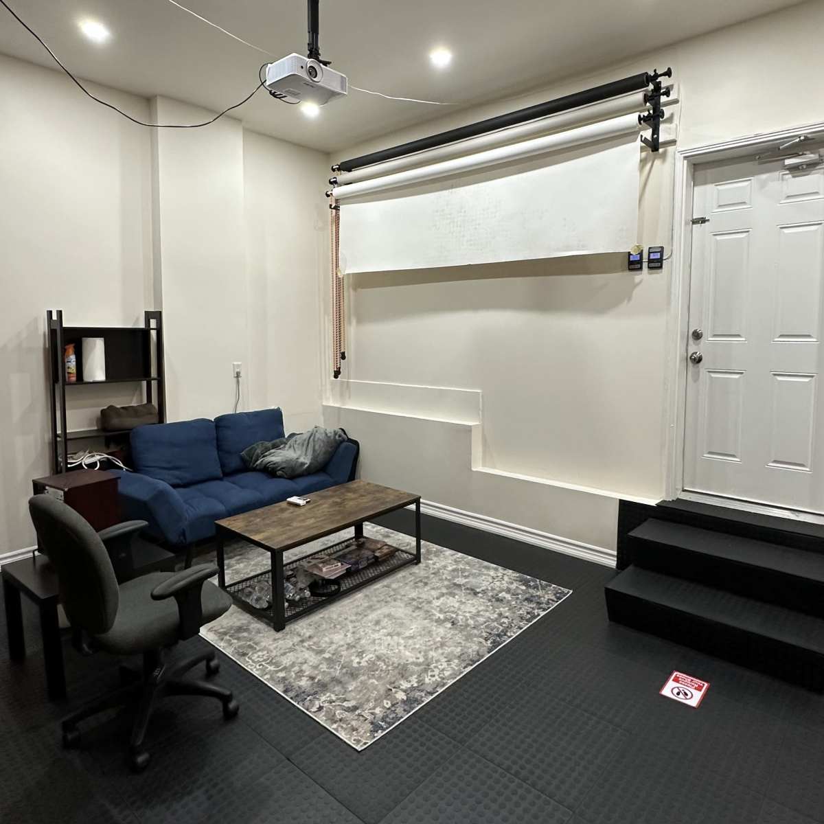 A small room with a blue couch, a wooden coffee table, an office chair, a door, and a projection screen mounted on the wall.