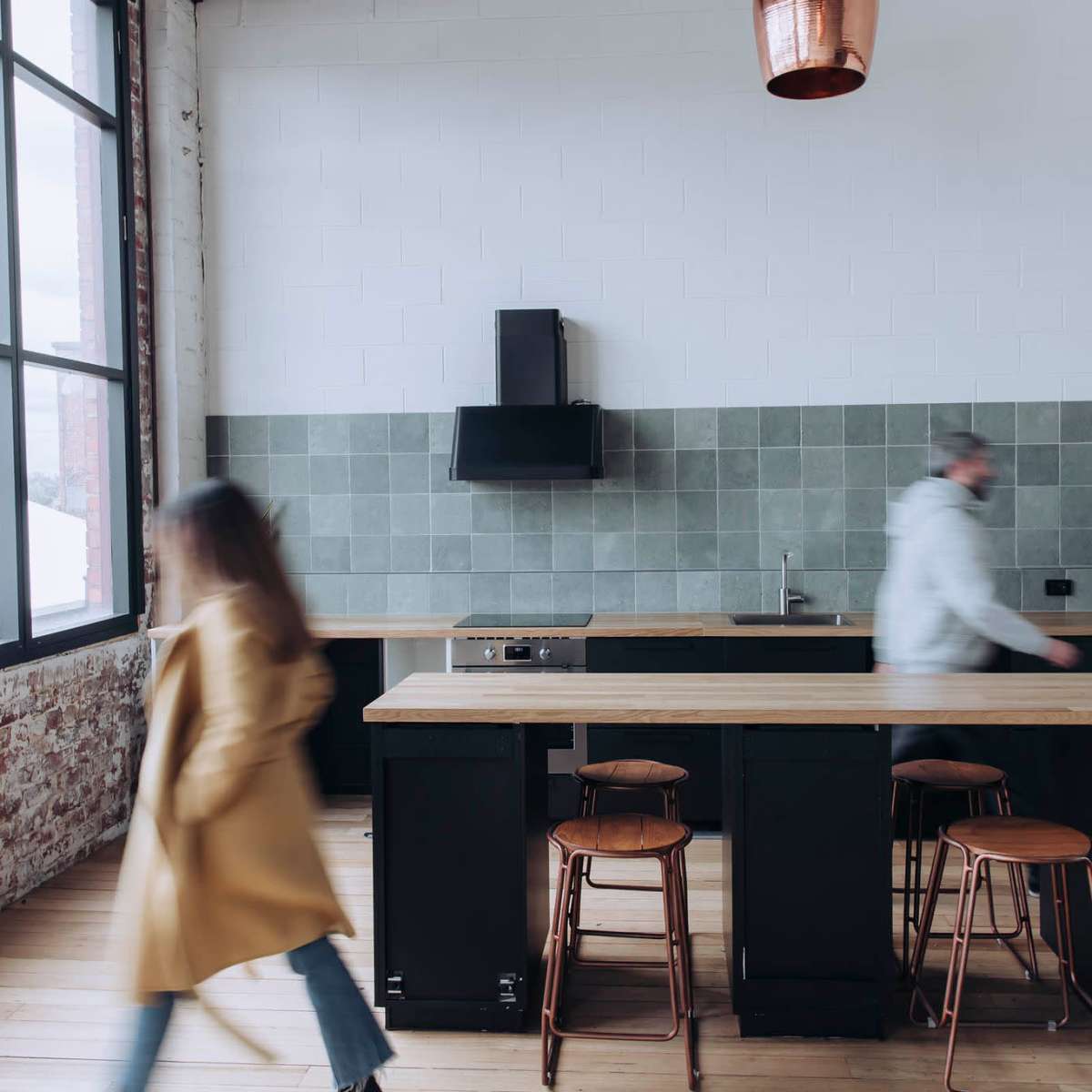 A kitchen features a large countertop with three wooden stools, a black refrigerator, and a person walking past while another stands at the counter.