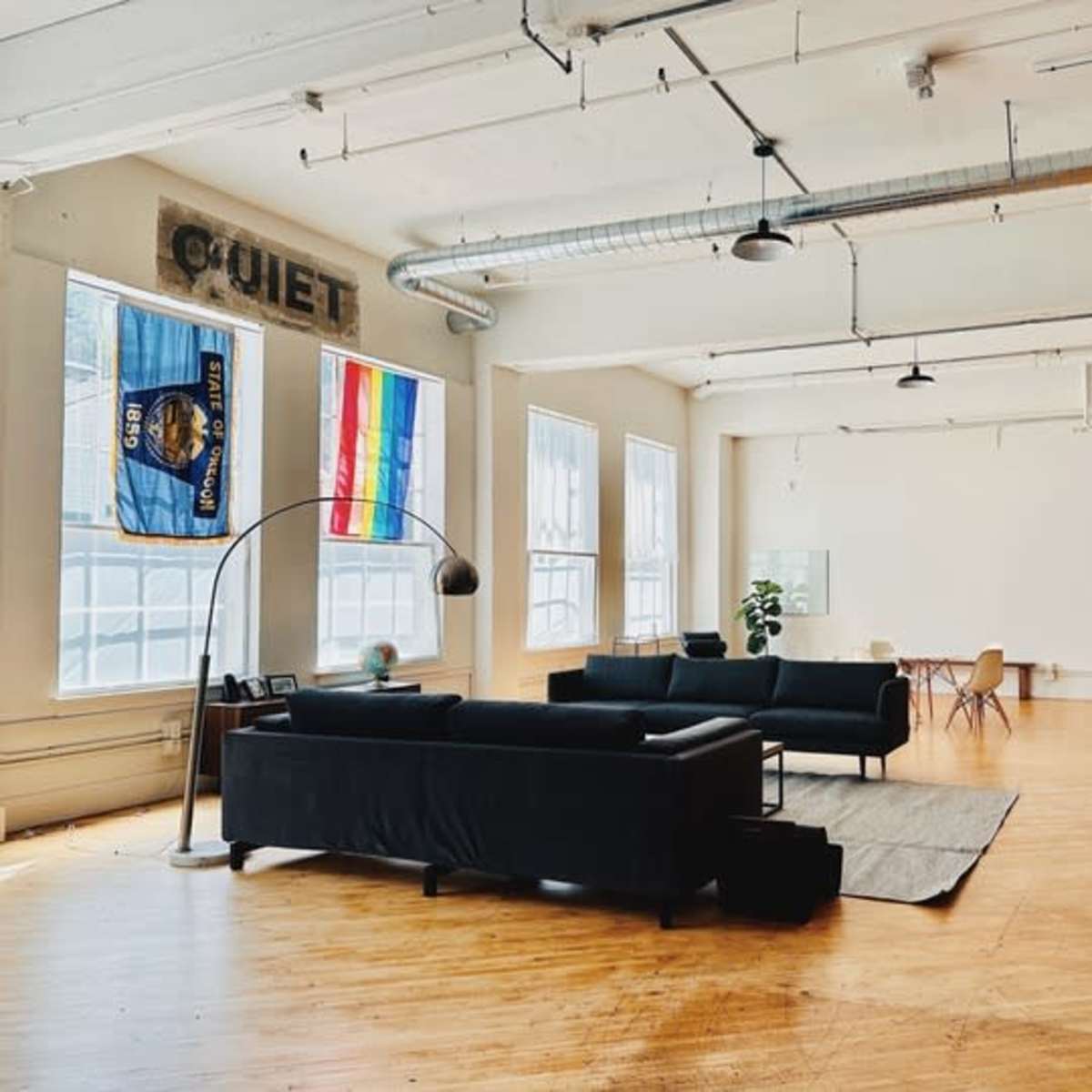 A spacious, minimally furnished room features a black sofa, a large window with a rainbow flag, and a dining area with a wooden table and chairs.