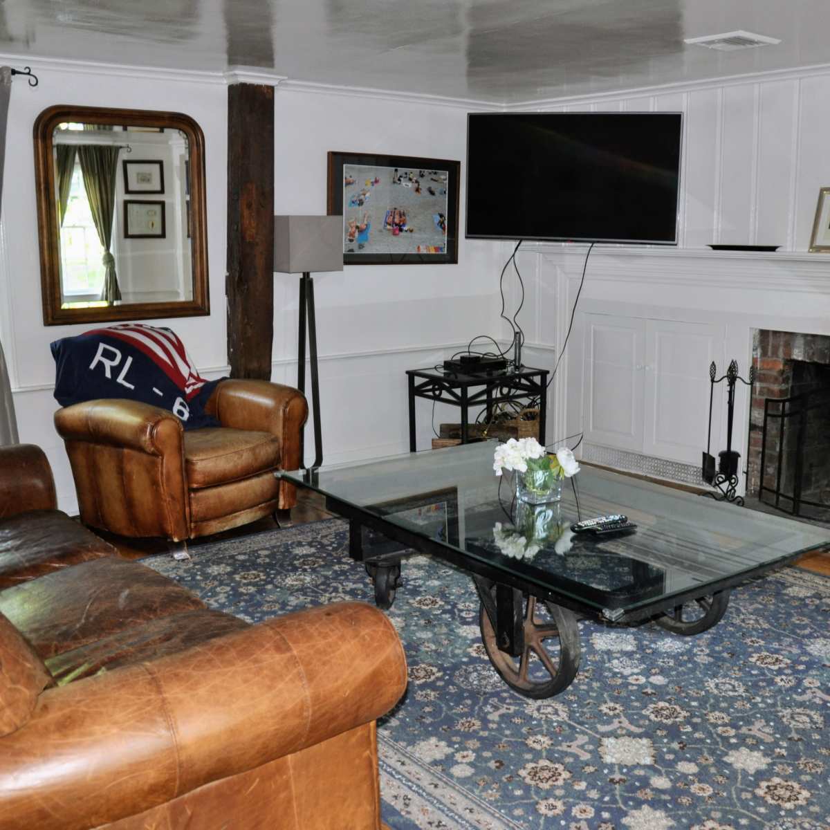 The living room features a leather chair, a glass coffee table on a patterned rug, and a television mounted above a white brick fireplace.
