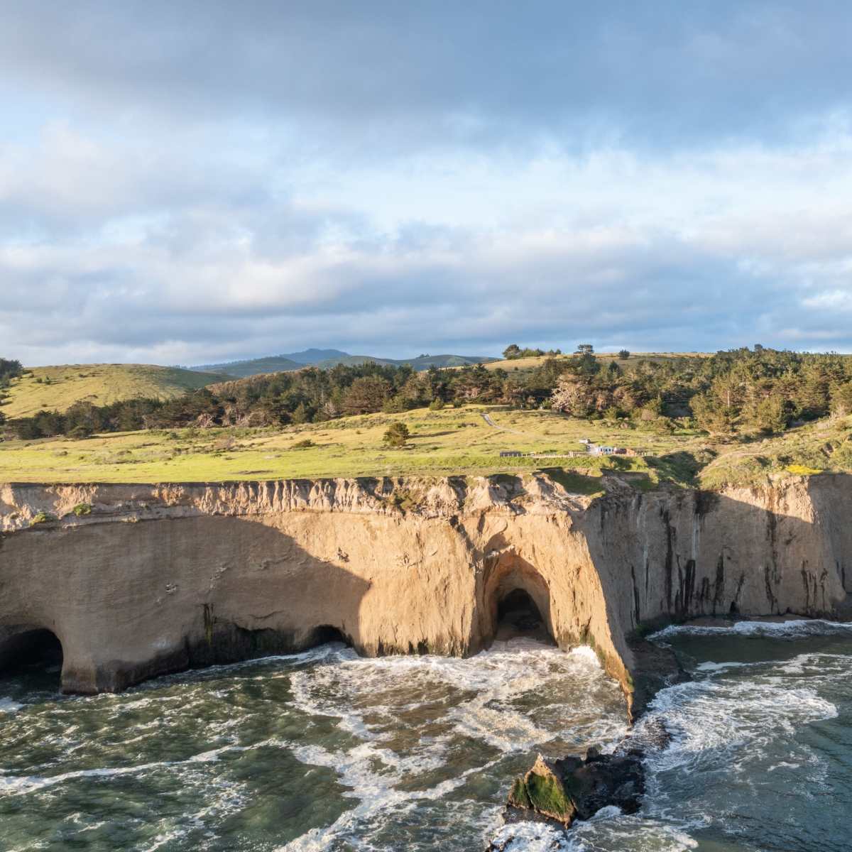 A rugged coastline with cliffs meeting the ocean, framed by green hills and patches of clouds in the sky.
