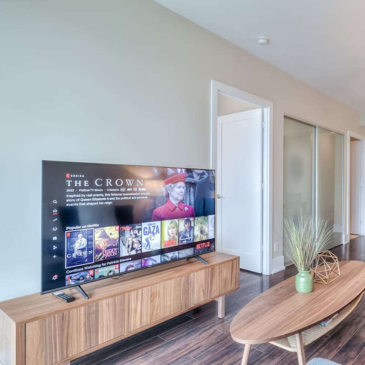 A modern living area features a television mounted on a wooden stand, a blue chair, and a dining area with a table and chairs, illuminated by overhead lighting.