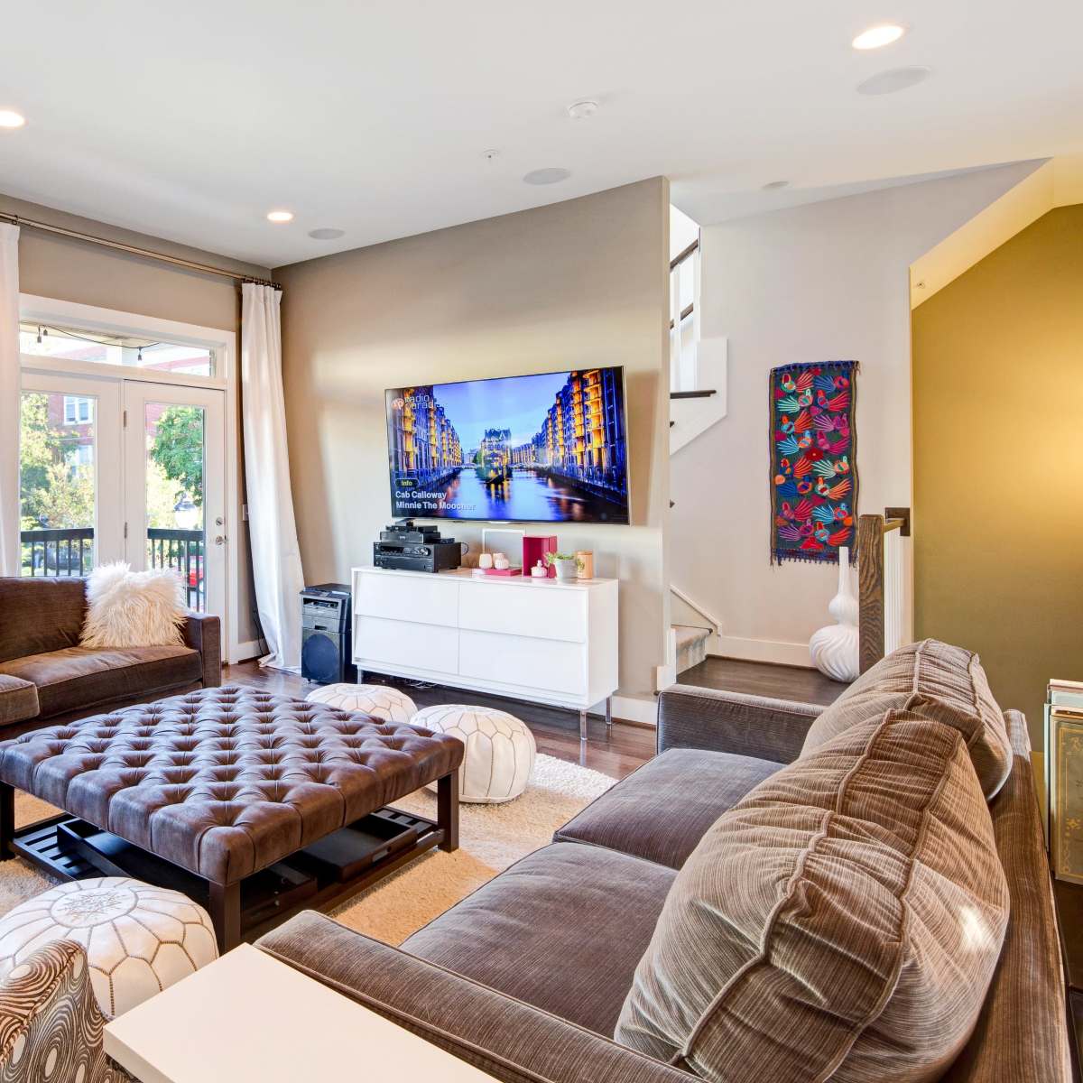The living room features a large sectional sofa, a coffee table, and a television mounted on the wall, illuminated by natural light from large windows.