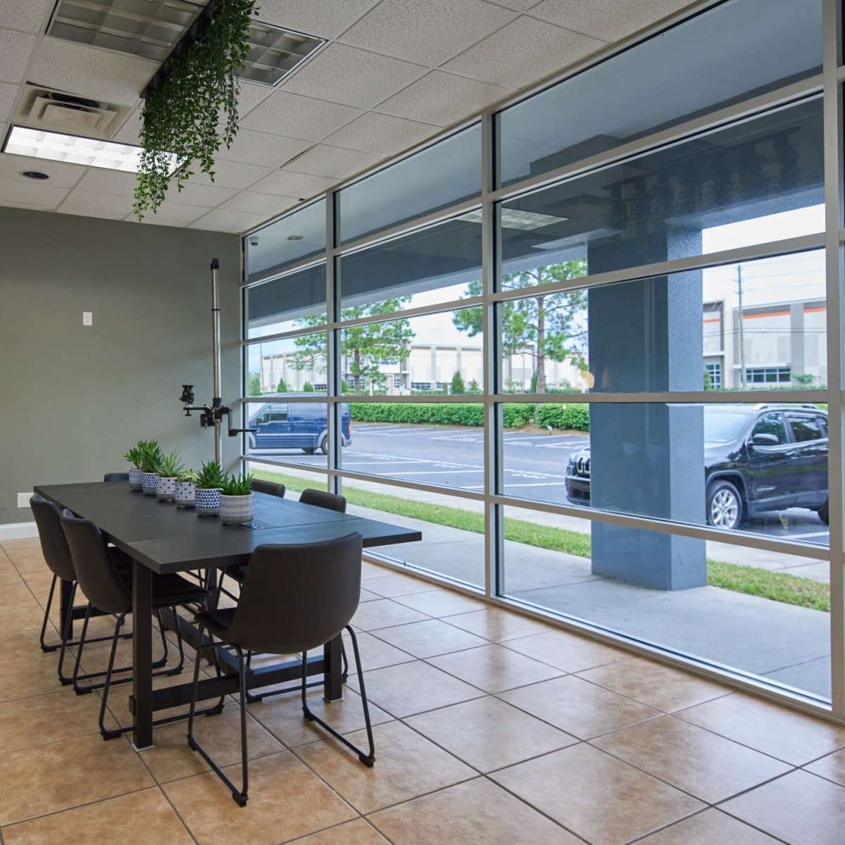 A modern meeting room features a large table with chairs, surrounded by windows offering a view of an outdoor parking area.