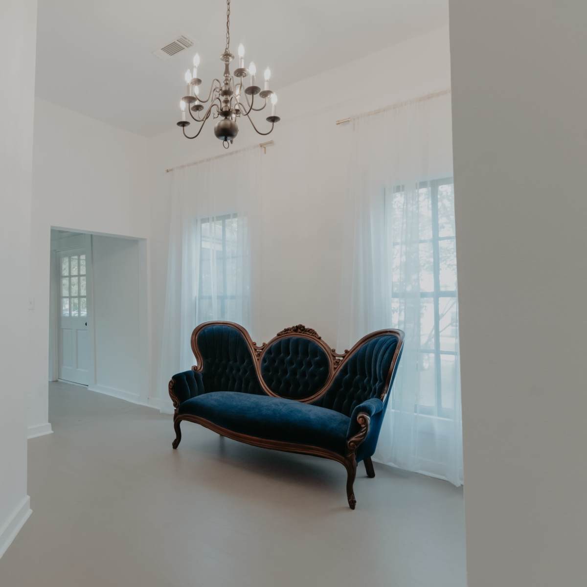The scene features a spacious room with a blue velvet sofa, a chandelier, and large windows dressed with sheer curtains.