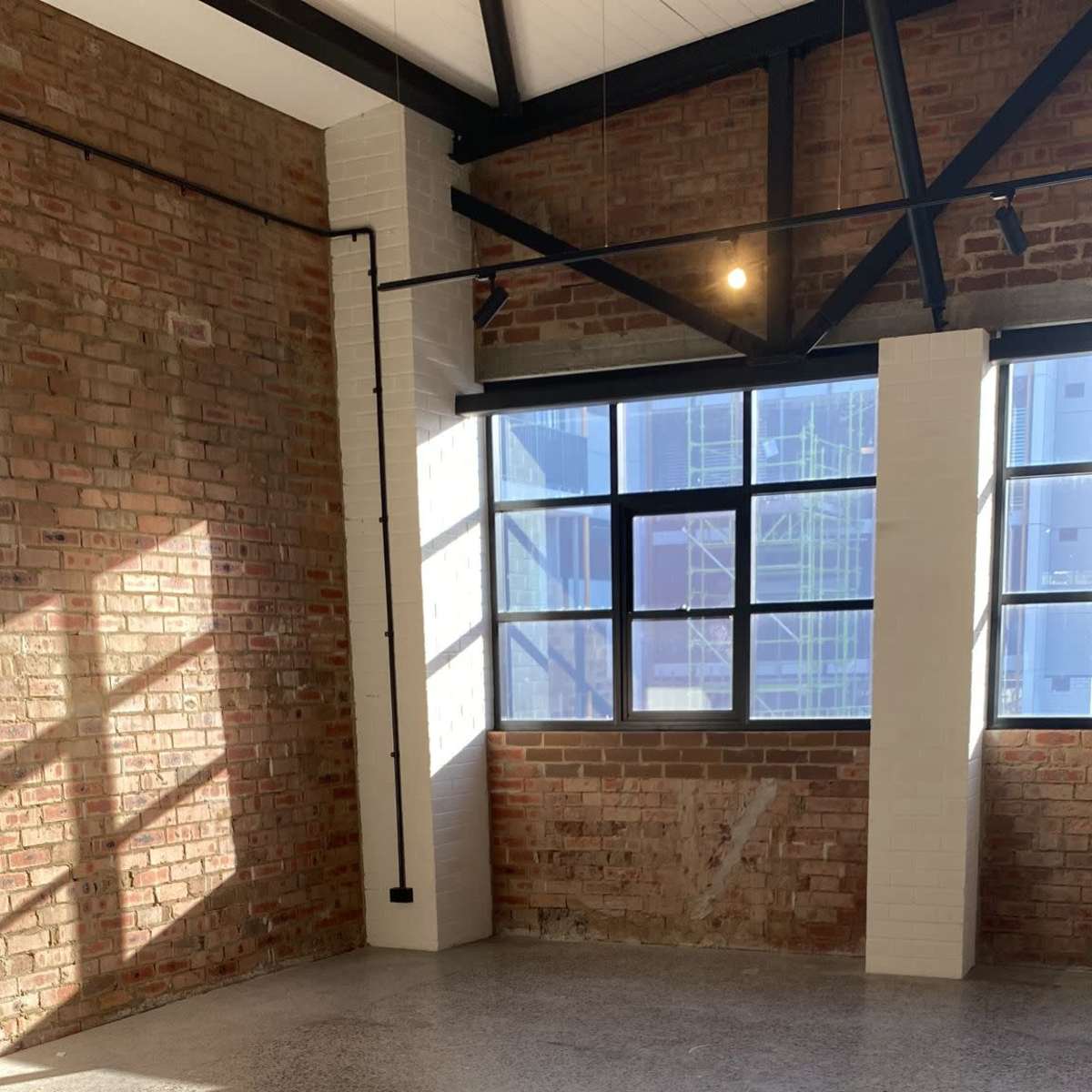 A spacious room with exposed brick walls and large windows, featuring a concrete floor with light reflecting on it.