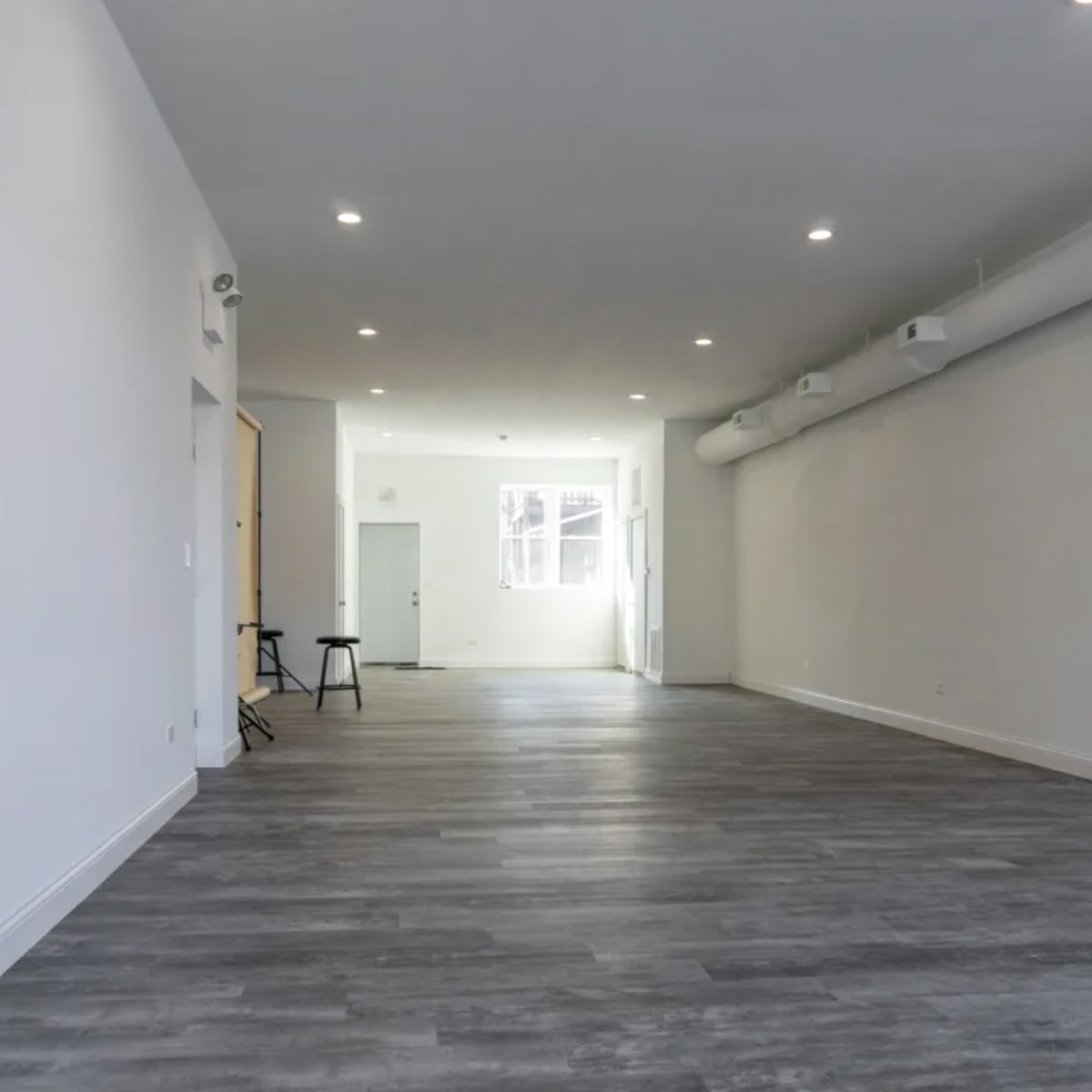 A spacious, empty room with grey flooring and white walls, featuring overhead lighting and windows at one end.